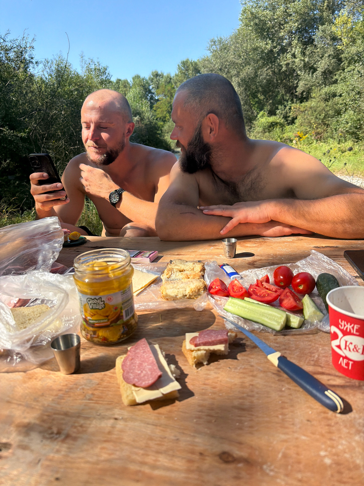 Two shirtless adult males are seated at a wooden picnic table outdoors. The male on the left is holding a smartphone and looking at it. He has a bald head and a beard. The male on the right has a beard and his arms are crossed on the table. The table is set with food items including a jar of pickles, sliced tomatoes, sliced cucumber, bread, cheese, and salami. There is also a red paper cup with Cyrillic writing on it. The background shows lush green trees and a clear blue sky, suggesting a natural, possibly rural or park-like setting. The presence of the red cup with Cyrillic writing, specifically "УЖЕ K&R ЛЕТ", which translates to "Already K&R Summer", indicates a connection to a specific event or brand, potentially related to Yessentuki, Russia, a well-known resort town in the Stavropol Krai region of Russia famous for its mineral waters and sanatoriums. The overall scene depicts a casual outdoor meal.