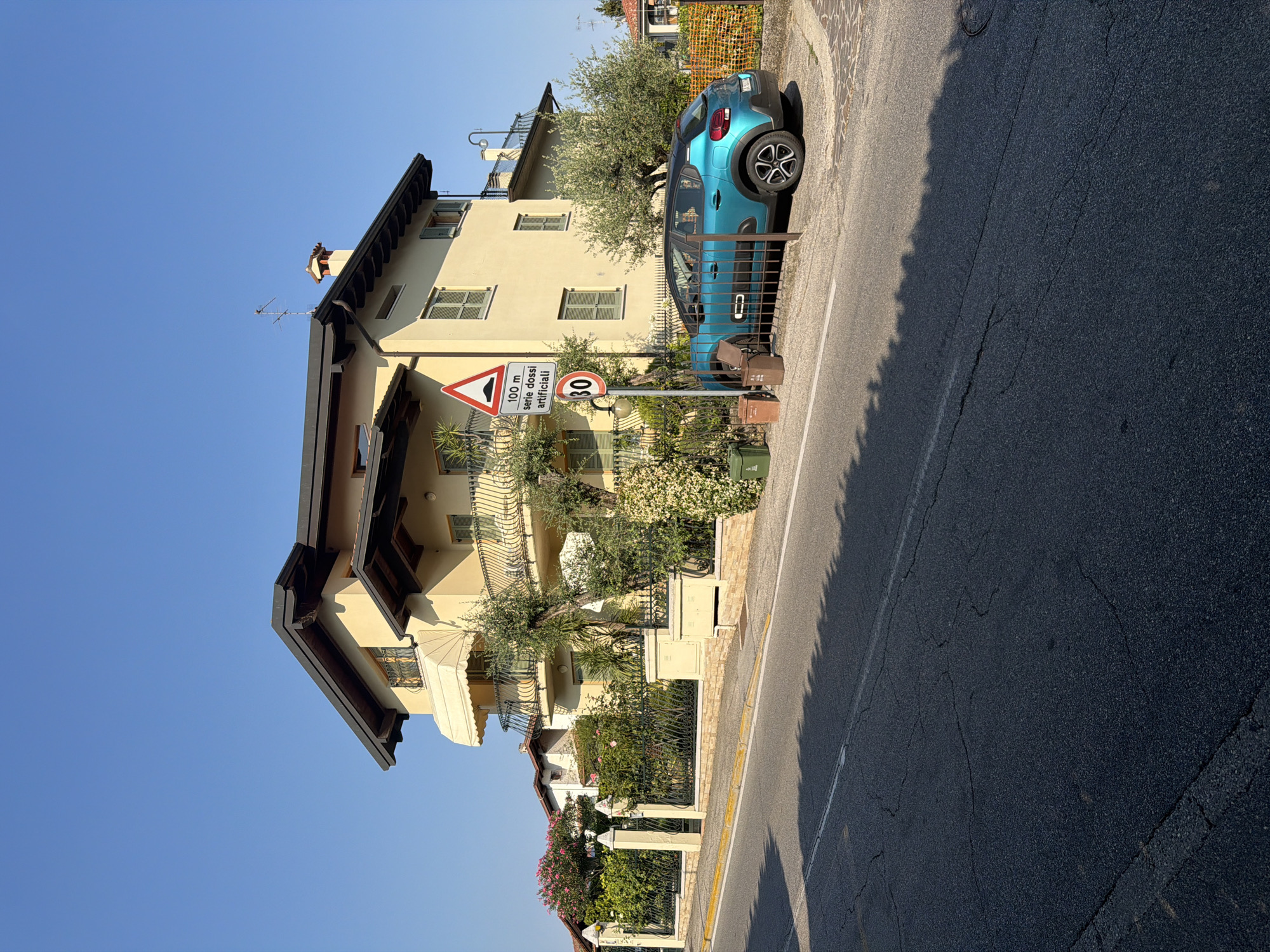 The image depicts a bright, sunny day in Padenghe sul Garda, Italy, with a clear blue sky dominating the upper portion. In the foreground, a paved road with visible cracks and a white line runs horizontally across the frame. A dark shadow, cast by foliage, stretches across the road from the left side.
On the right side of the road, a light yellow, multi-story house with a dark-tiled roof and balconies is the central architectural feature. The house has green shutters on some windows. Adjacent to the house, a turquoise hatchback car is parked. Beside the car, a metal fence encloses a small area, and to its left, a traffic sign with a red border indicates "100 m serie dossi artificiali" (100 m series of artificial speed bumps) and a speed limit of 30. Two trash bins, one green and one brown, are positioned near the fence.
Behind the house, a lush garden with various plants and trees, including olive trees, is visible. A black metal fence borders the property, separating it from the road. In the background, other houses and vegetation can be seen, with an orange net fence to the far right. The overall scene suggests a residential street in a town or village.