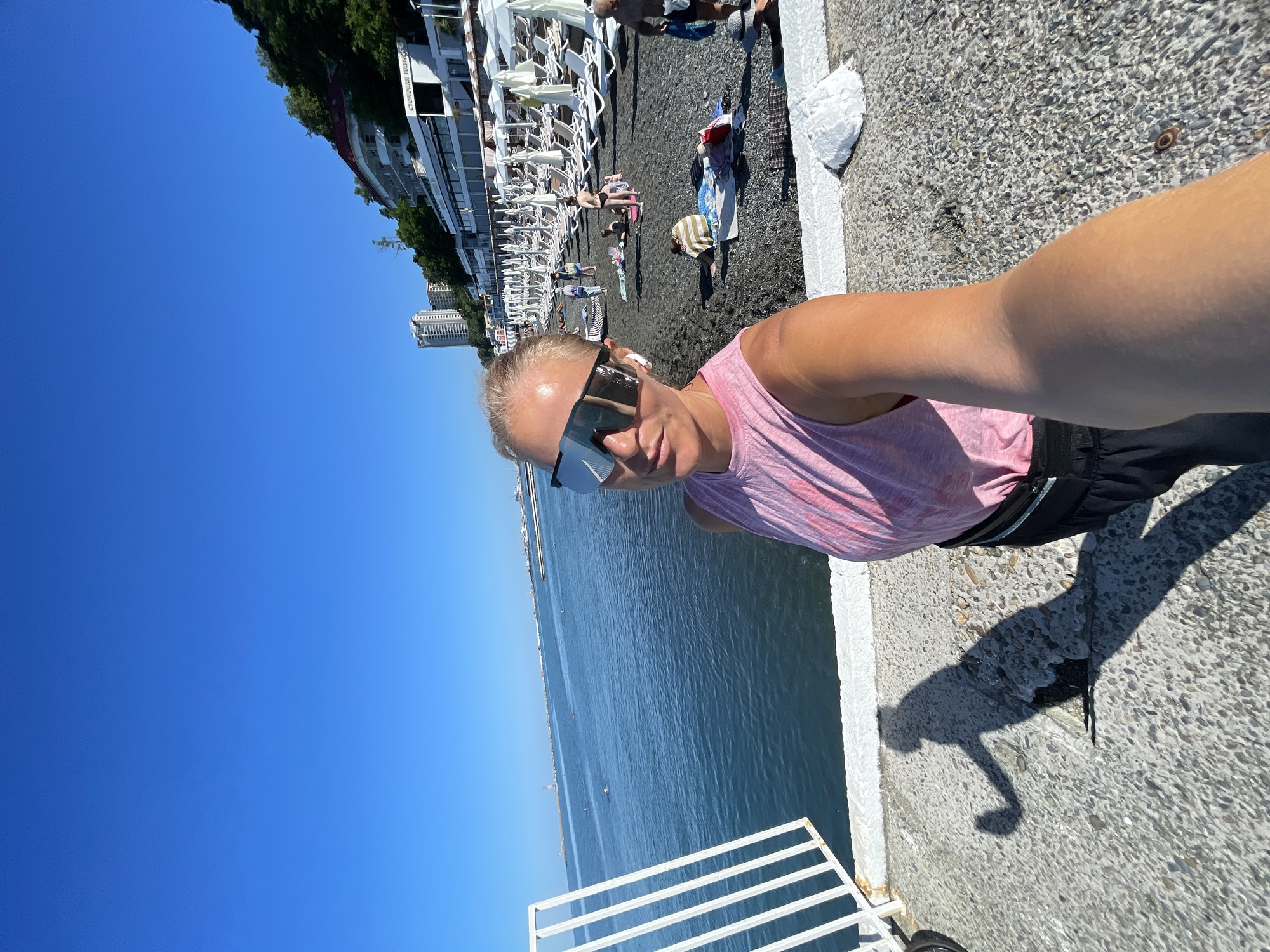 A woman with blonde hair pulled back, wearing large, reflective sunglasses, a pink tank top, and dark shorts, takes a selfie on a sunny day at the beach in Sochi, Russia. She is standing on a concrete walkway next to a white railing, with the dark pebble beach and the blue Black Sea stretching out behind her. A line of white beach umbrellas and lounge chairs are visible on the beach, along with several other beachgoers. In the background, buildings and lush green hillsides rise up from the coast. The sun casts a long shadow of the woman onto the walkway.