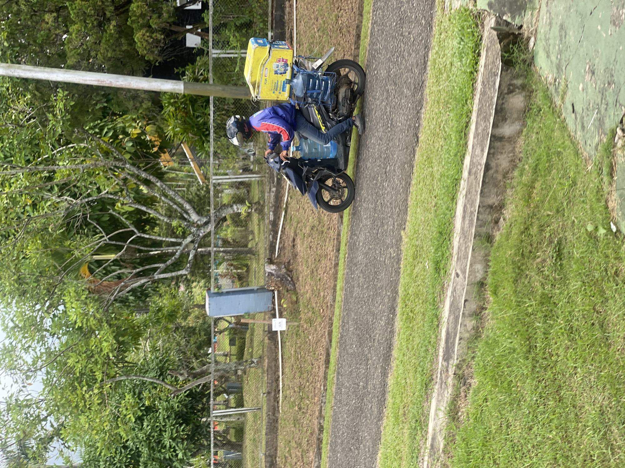 A person on a scooter drives on a paved road through a green, outdoor area. The person is wearing a blue jacket with red accents, blue jeans, and a black helmet. The scooter is dark blue and has a yellow cooler box strapped to the back. Two large water bottles are attached to the sides of the scooter. The area around the road is grassy with scattered trees and a fence. A tall, grey pole stands to the right of the frame. The sky is overcast. The image was taken in Banjarbaru, Indonesia.
