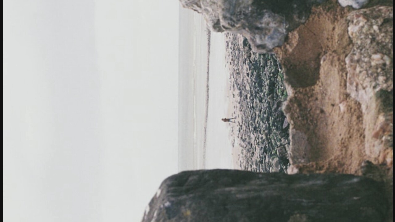 In the city of Unknown, Unknown, a person wearing an orange jacket stands on a rocky beach, facing the ocean. The person is viewed from behind, and a small, dark dog stands near their feet. The beach is covered in smooth, grey stones. In the foreground, large, dark rocks partially obscure the view of the beach and ocean. The sky is overcast, with a pale grey hue. The ocean stretches to the horizon, with a faint line of what appears to be a breakwater or submerged structure visible in the distance.