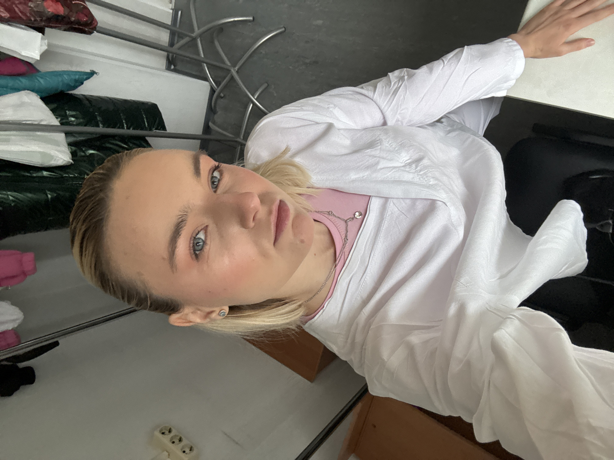 A medium shot captures a fair-skinned person with blonde hair, wearing a light pink top and a white button-down shirt or lab coat. The person is positioned at a slight angle. The person has light eyes, a light complexion, and is wearing a necklace. The subject is at a table with a chair and some miscellaneous items. The location cannot be determined.