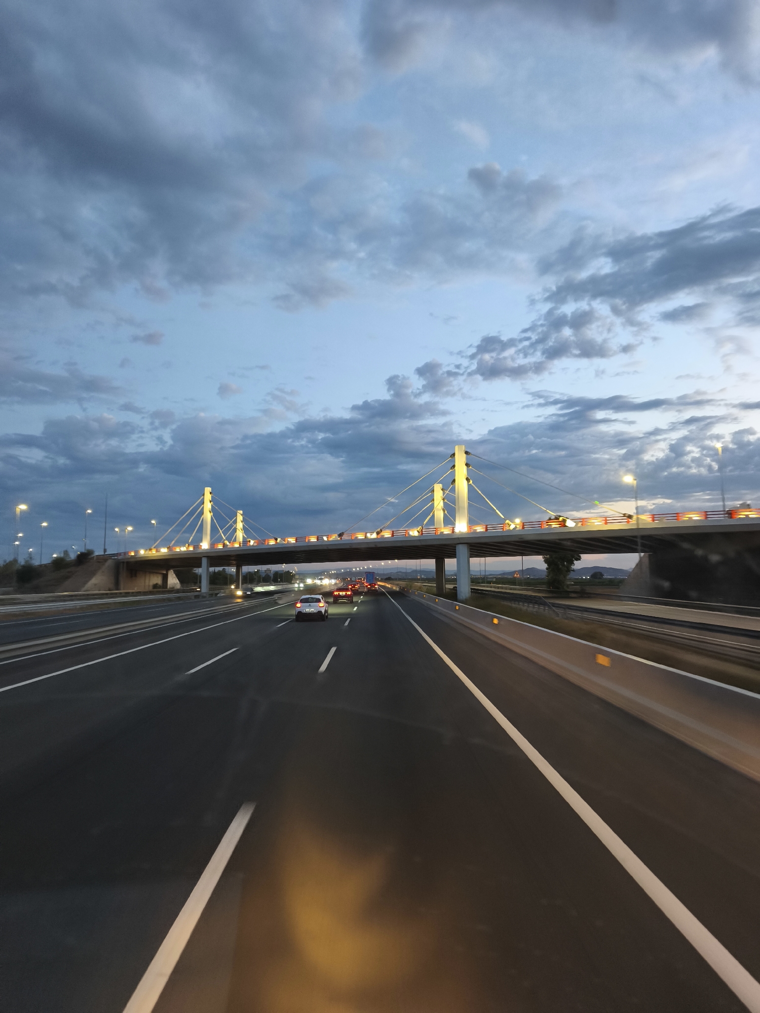 The image depicts a highway at dusk, with a suspension bridge illuminated by golden lights spanning across it. Several cars are visible on the highway, indicating ongoing traffic. The sky above is filled with dramatic, dark clouds, suggesting an impending storm or the aftermath of one. Streetlights line the highway, casting a soft glow on the road. The scene appears to be in the city of Chiva, Spain, given the reference to the location. The overall atmosphere is serene yet dynamic, with the contrast between the dark sky, the illuminated bridge, and the moving vehicles.