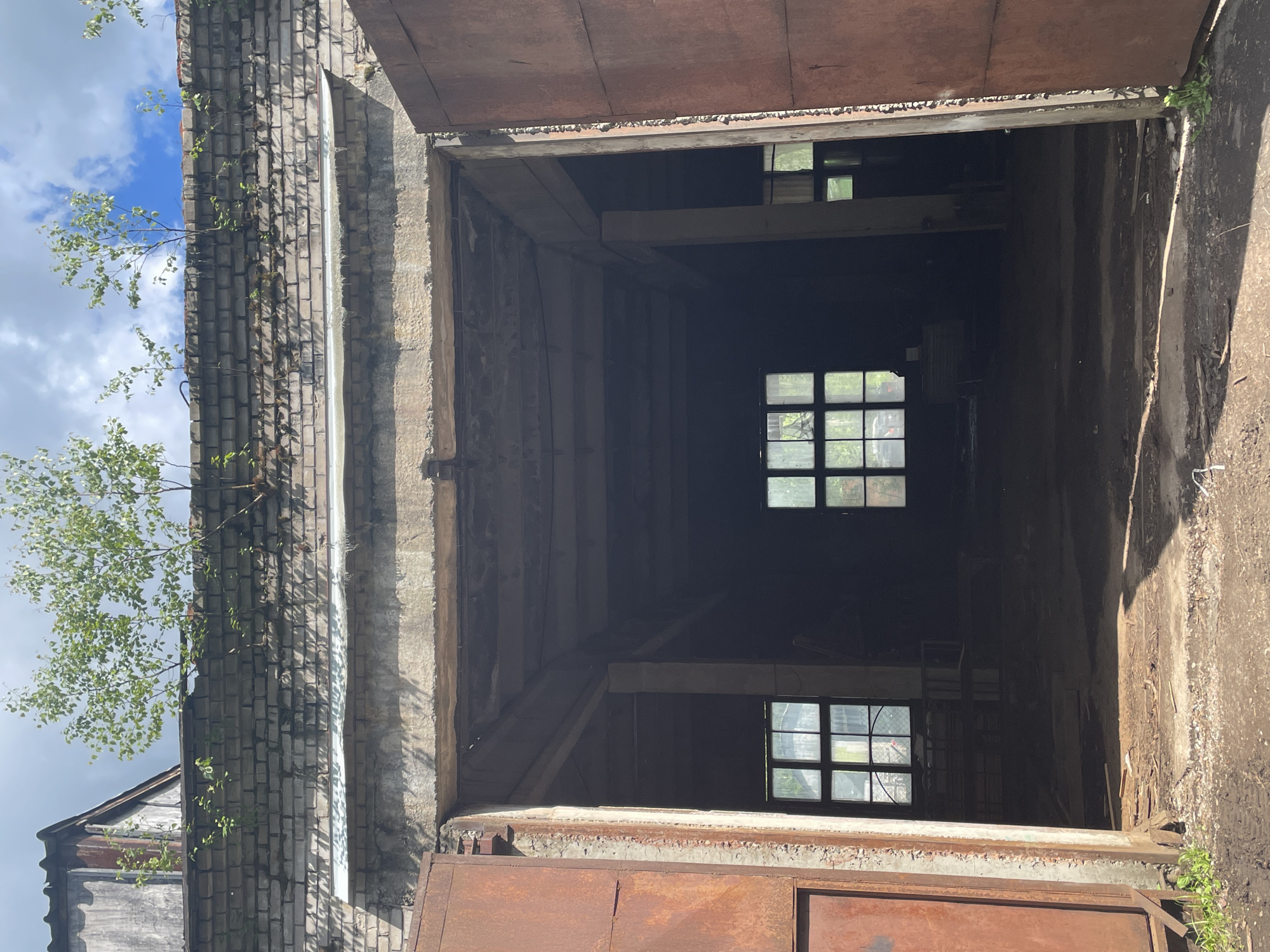 The image shows the interior of an abandoned industrial building. The main subject is a large, open doorway leading into a dark space. The doorway is flanked by two large, rusty metal doors, one of which is partially open. Inside, there are two windows visible, with panes of glass, and what appears to be scattered debris on the floor. Trees are growing on top of the brick wall above the doorway. The overall scene suggests neglect and disuse. The location is not identifiable within the city of Saint Petersburg, Russia.