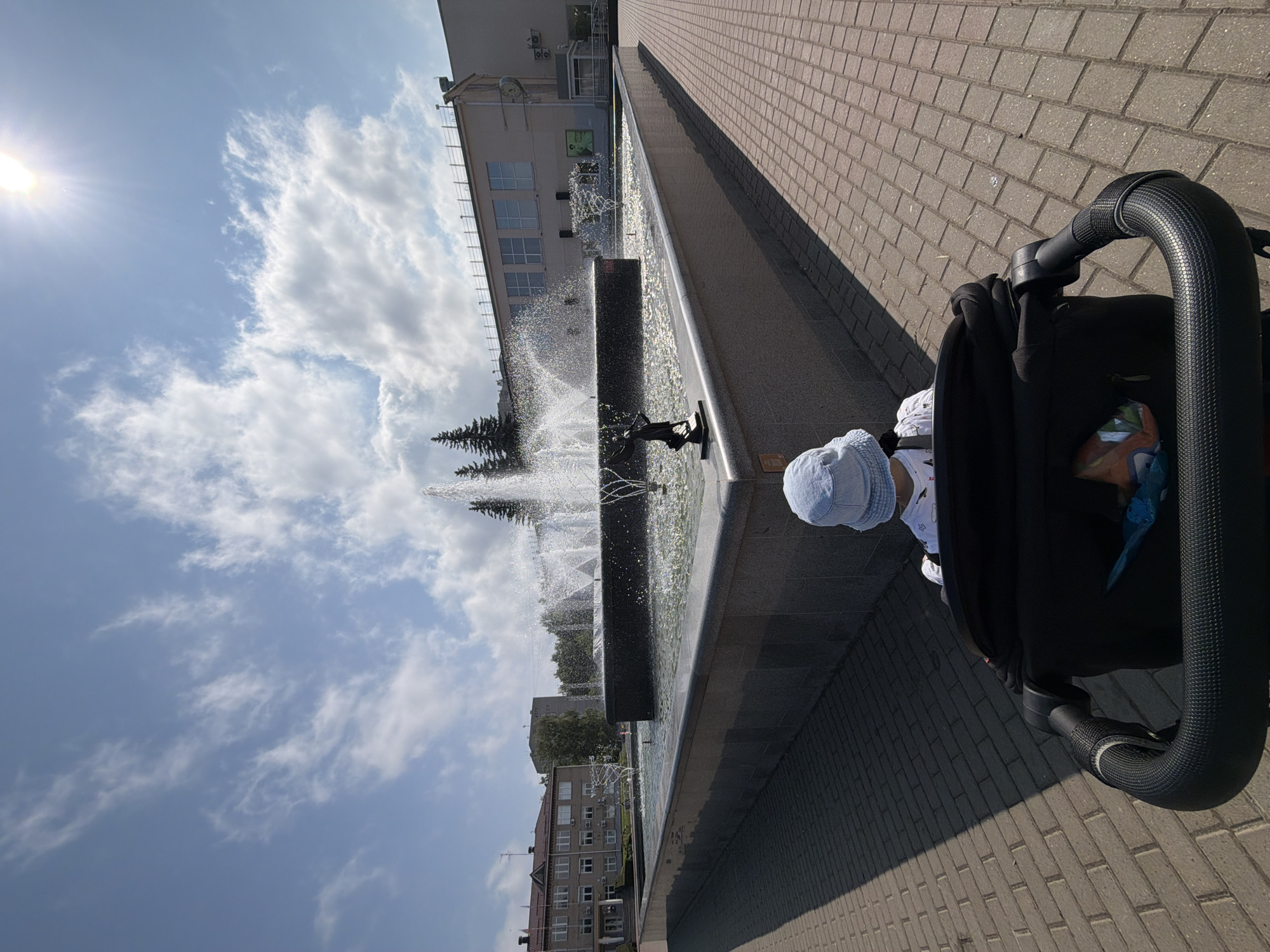 An overhead shot captures a child in a stroller facing a fountain in Chaikovsky, Russia. The fountain is central, with water jetting upwards. The child is wearing a light blue bucket hat and a white shirt with a design. The stroller is black, and part of its handle is visible in the foreground. The fountain is surrounded by a stone plaza and a low stone wall. In the background, several buildings and trees can be seen under a bright, cloudy sky with the sun shining.