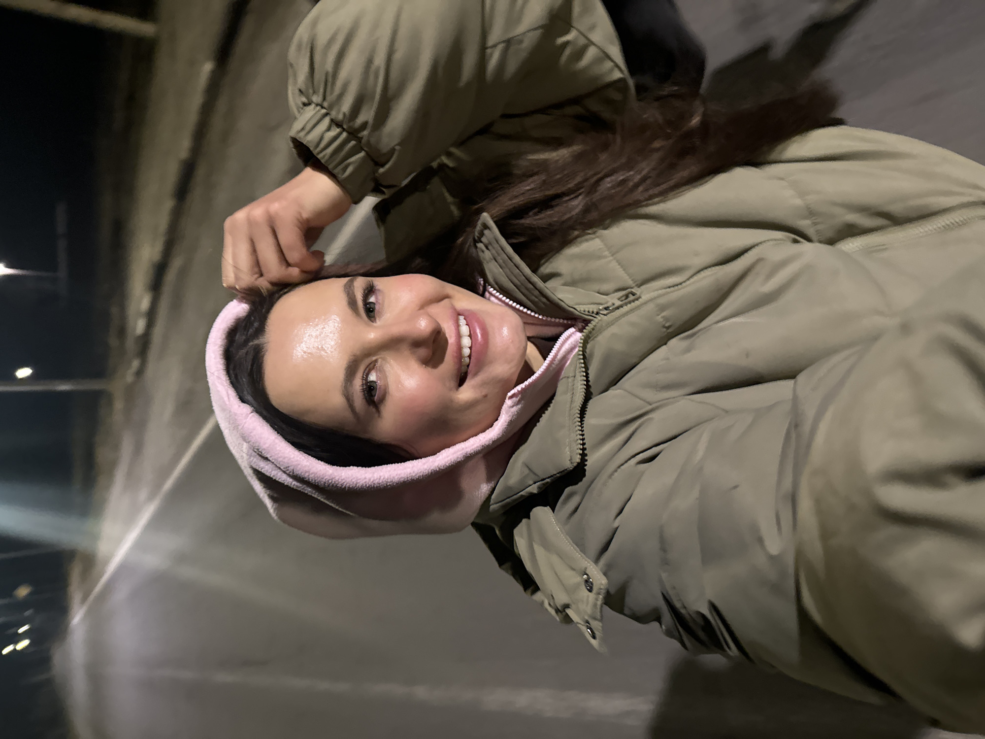 A woman wearing a khaki puffer jacket with a pink hood is smiling at the camera. She is standing on a paved surface. In the background is a concrete wall and what appears to be streetlights. The location is the city of Красный Маяк, Russia.