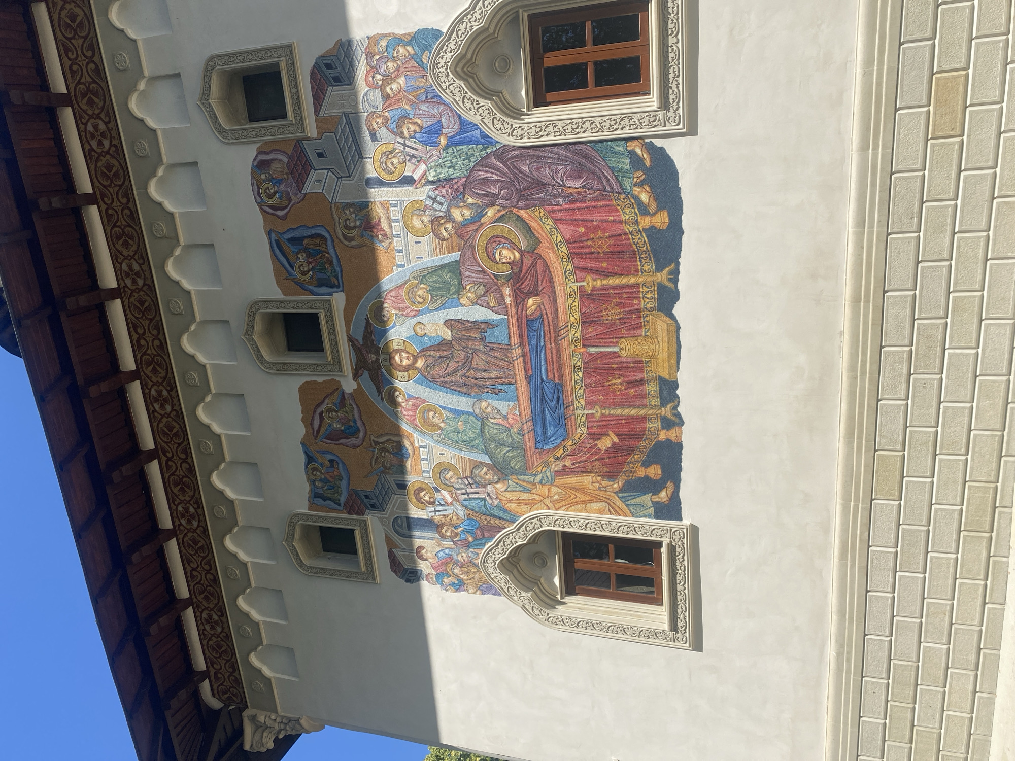 The image depicts a fresco on the exterior wall of a building, likely a church or monastery, located in Filioara, Romania. The fresco portrays a religious scene with numerous figures. In the center, a deceased figure lies on a raised bed covered with a red and gold patterned cloth, surrounded by other individuals who appear to be mourning or paying respects. Above them, celestial beings or angels are depicted. The fresco is rich in color and detail, with figures rendered in traditional Byzantine or Orthodox style. The building itself is white-plastered with ornate architectural details, including arched window frames with intricate carvings and a dark wooden roof with decorative trim. There are also two visible windows with dark wooden frames. The lower portion of the wall is constructed of light-colored brickwork. The sky above is a clear, bright blue, indicating it is a sunny day. The overall scene suggests a historical or religious context, typical of Orthodox Christian architecture found in Romania.