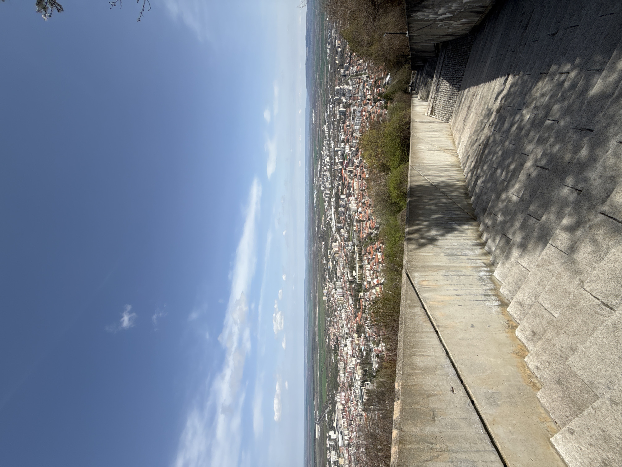 The image shows a view from the Monument to 1300 Years of Bulgaria in Shumen, Bulgaria. The vantage point offers an aerial view of the city. The right side of the image features the edge of a gray concrete structure with visible steps leading down. Tree shadows are cast on the steps. To the left of the structure, the cityscape unfolds, densely packed with buildings. Beyond the city, a flat landscape stretches to the horizon, partly obscured by atmospheric haze. The sky occupies the left side of the image, showing a gradient of blue with scattered white clouds.