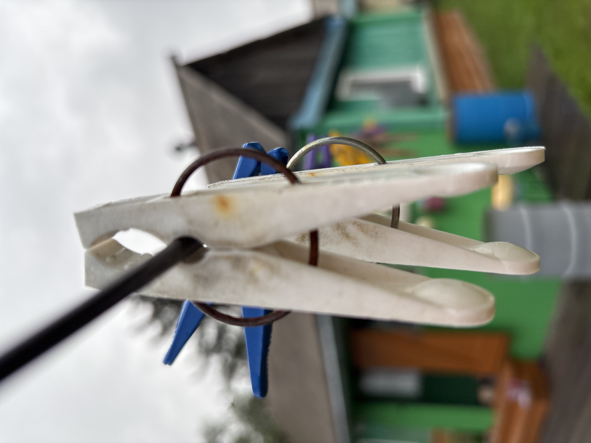 The image is a close-up, shallow depth of field shot of two white plastic clothespins on a dark clothesline. The clothespins are positioned vertically and appear to be slightly dirty, with some rust spots visible on one. Behind the clothespins, a blurry green house with a dark grey roof and a wooden porch is visible. A blue barrel and a grey cylinder are also present in the background. The sky is overcast and grey. The scene is set in an outdoor environment, likely a backyard, with green grass visible in the foreground and background. The location is specified as Gonja, Russia. No people or actions are depicted.