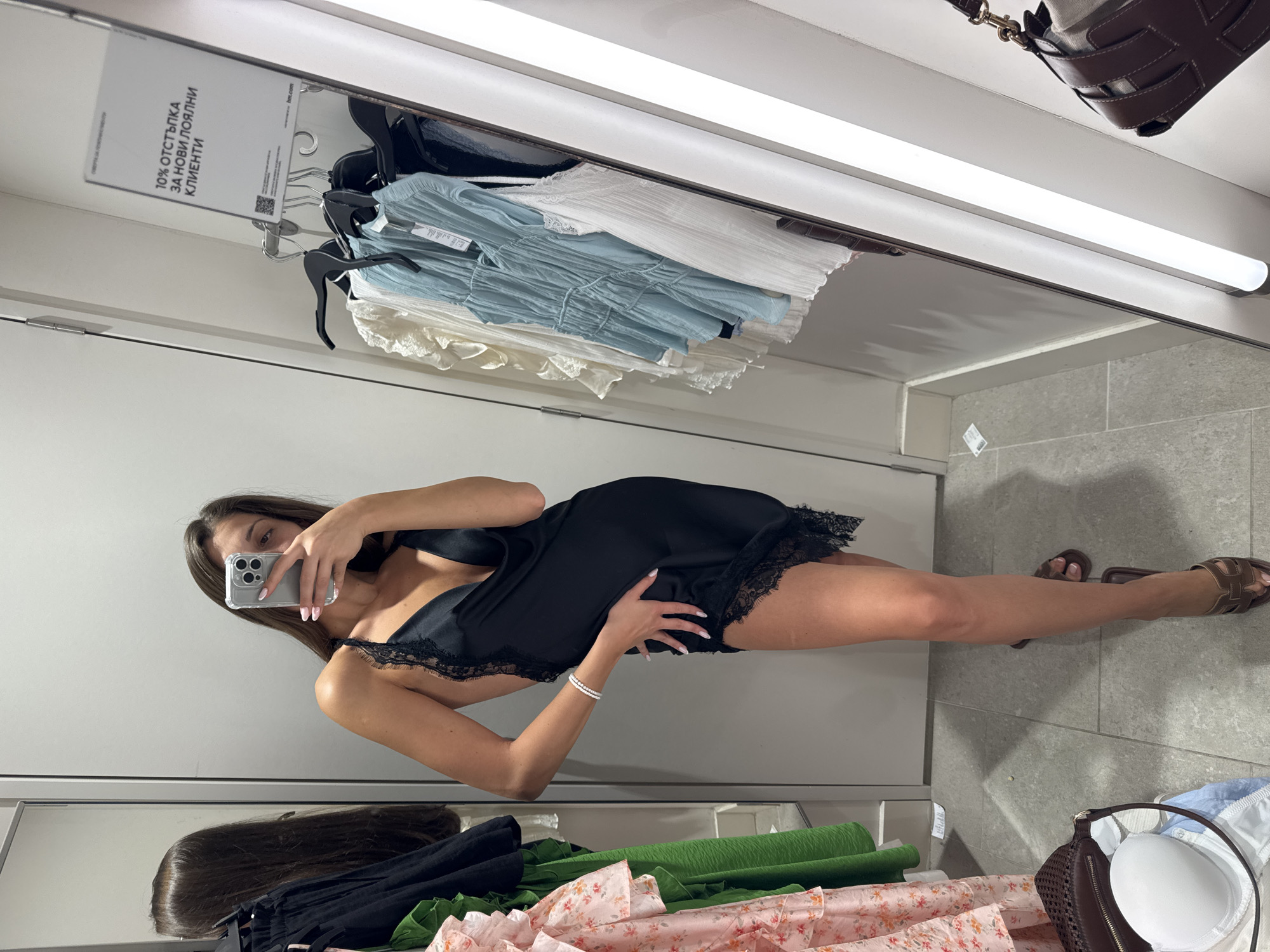 The image captures a mirror selfie taken by a young woman in what appears to be a fitting room or dressing room of a retail clothing store in Burgas, Bulgaria.
The central subject is a woman with long, dark brown hair, seen from the waist up in her reflection and full body in the mirror. She is wearing a black slip dress with delicate black lace trim along the neckline and hem. She has light-colored nail polish on her fingernails and a white beaded bracelet on her left wrist. Her face is partially obscured by a silver-colored smartphone, which she holds in her right hand, actively taking the mirror selfie. She is wearing brown flat sandals on her feet.
The setting is an indoor space with white or light gray walls and light gray tiled flooring. A long fluorescent light fixture is visible on the ceiling or above the mirror. In the background, behind the woman's reflection, there is a clothes rail with various garments, including light blue ruffled tops/dresses and white blouses. Below the mirror, on a shelf or bench, are piles of clothing items, including green fabric, a pink floral patterned fabric, and other dark garments. On the floor near her feet, a brown woven bag is visible, partially open, revealing a white padded bra cup inside.
A white sign is hanging above the clothes rail, displaying text in Cyrillic which reads: "10% ОТСТЪПКА ЗА НОВИ ЛОЯЛНИ КЛИЕНТИ". This translates to "10% DISCOUNT FOR NEW LOYAL CLIENTS". The sign also features a QR code and a small "hm.com" logo, suggesting the location is likely an H&M store. The activity depicted is the woman trying on clothing and documenting her appearance with a selfie.