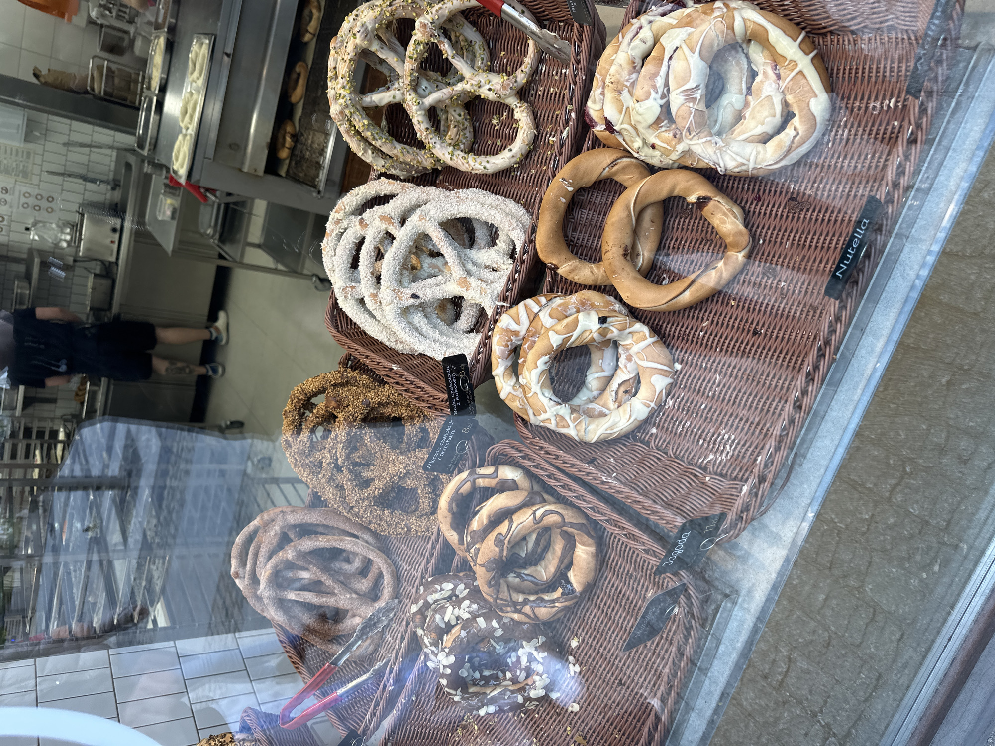 The image shows a display case filled with a variety of pretzels, likely in a bakery or cafe. The pretzels are arranged in wicker baskets, each with a different topping or flavor, including nuts, white chocolate with coconut, milk chocolate, and Nutella. Price tags accompany some of the pretzels. Through the glass of the display case, reflections of the interior of the bakery/cafe and a person are visible. Based on the price tag information, this display is located in Wrocław, Poland.