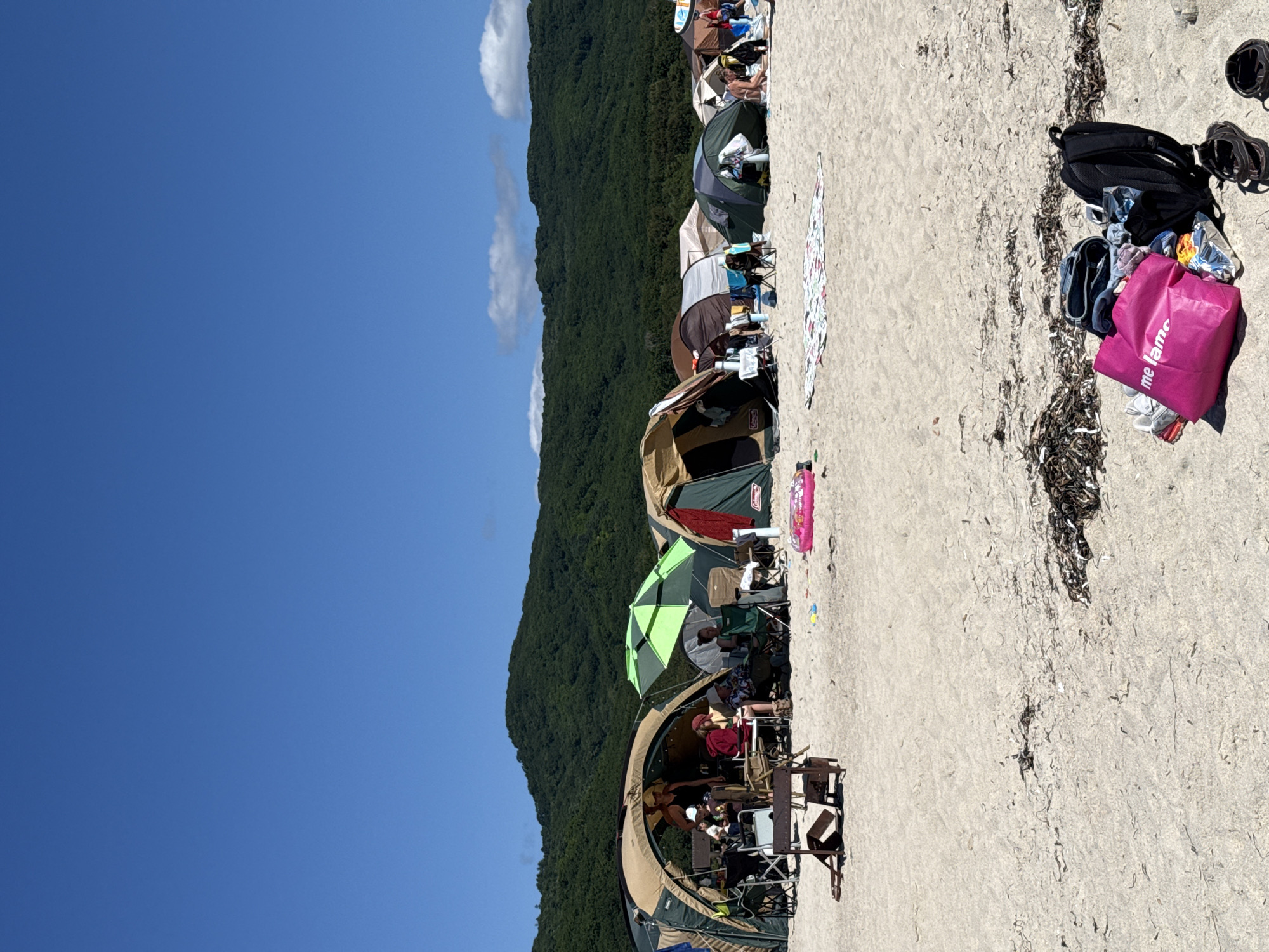 A group of people are camping on a sandy beach, likely in Nakhodka, Russia, given the lush green mountainous backdrop and the presence of tents and beach gear. The sky is a clear, bright blue with a few wispy clouds visible above the mountains. The beach is lined with several tents of various colors and sizes, some open and occupied, while others appear to be closed. People are seen sitting in chairs outside the tents, socializing, and relaxing. There are also camping chairs, a small grill, a pink inflatable ring, and beach towels spread out on the sand. In the foreground, a pile of belongings includes a pink tote bag with the text "me lamo" visible, a black backpack, and a pair of sandals. The environment is a typical beach setting, with sand covering the ground and a dense forest covering the hills in the distance. The overall impression is a sunny day spent camping by the sea.