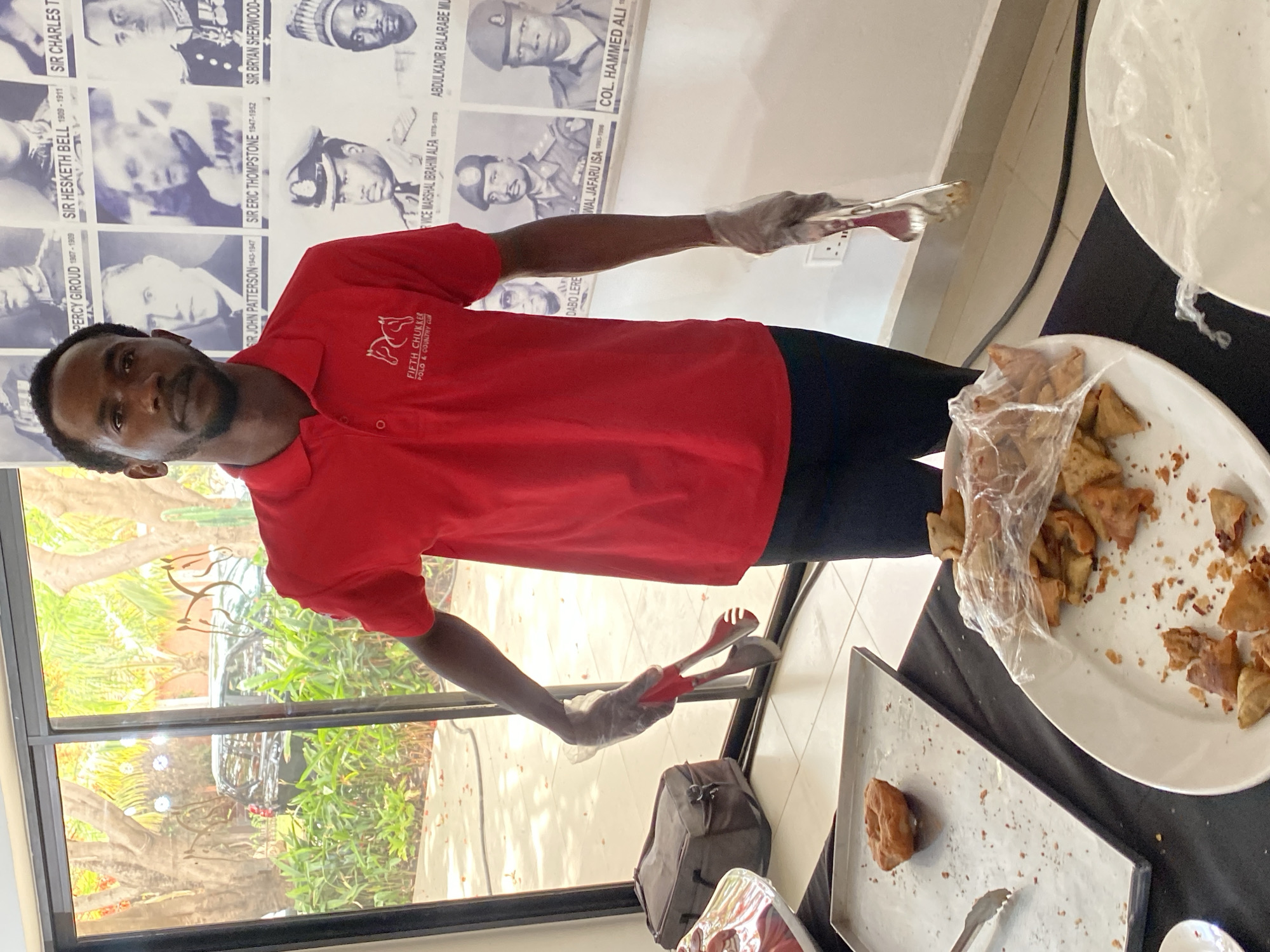 A man wearing a red polo shirt with the "Fifth Chukker Polo & Country Club" logo stands indoors. He wears clear plastic gloves and holds tongs. To his right is a plate of partially eaten samosas covered in plastic wrap, along with an empty metal tray with a single donut. Behind him are photos of people, including Percy Giroud, Sir Hesketh Bell, Sir Charles T., Sir John Patterson, Sir Eric Thompsonstone, Sir Bryan Sherwood, Abdulkadir Balarabe Mu, Col. Hammed Ali, Dabore Lere, and Wal Jafaru Isa. Through a window, exterior foliage and a vehicle are visible. The city of Kwarau, Nigeria, is not directly discernible from the image.