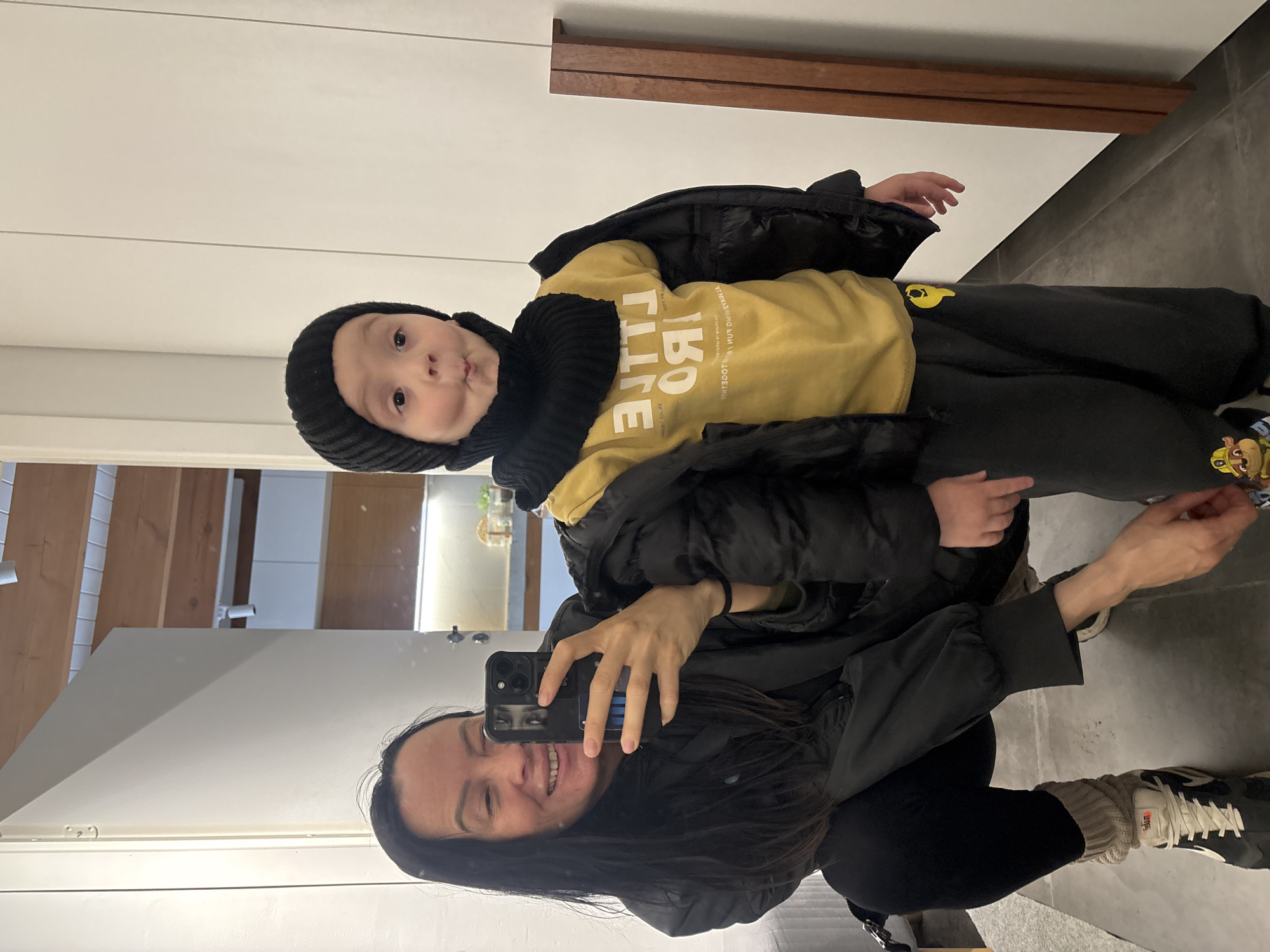 A woman is taking a selfie in a mirror with a young child. The woman has dark hair and is wearing a black jacket. She is holding a smartphone in her right hand. The child, wearing a black knit hat, a yellow sweatshirt with the word "LITTLE" printed in large white letters, and a black jacket, is being held by the woman. The background appears to be the interior of a home or apartment. There is a white cabinet behind the woman and child, and a partially visible entryway or room to the left. The floor is gray tile. The specific location in the city of Chaikovsky, Russia, cannot be determined from the image.