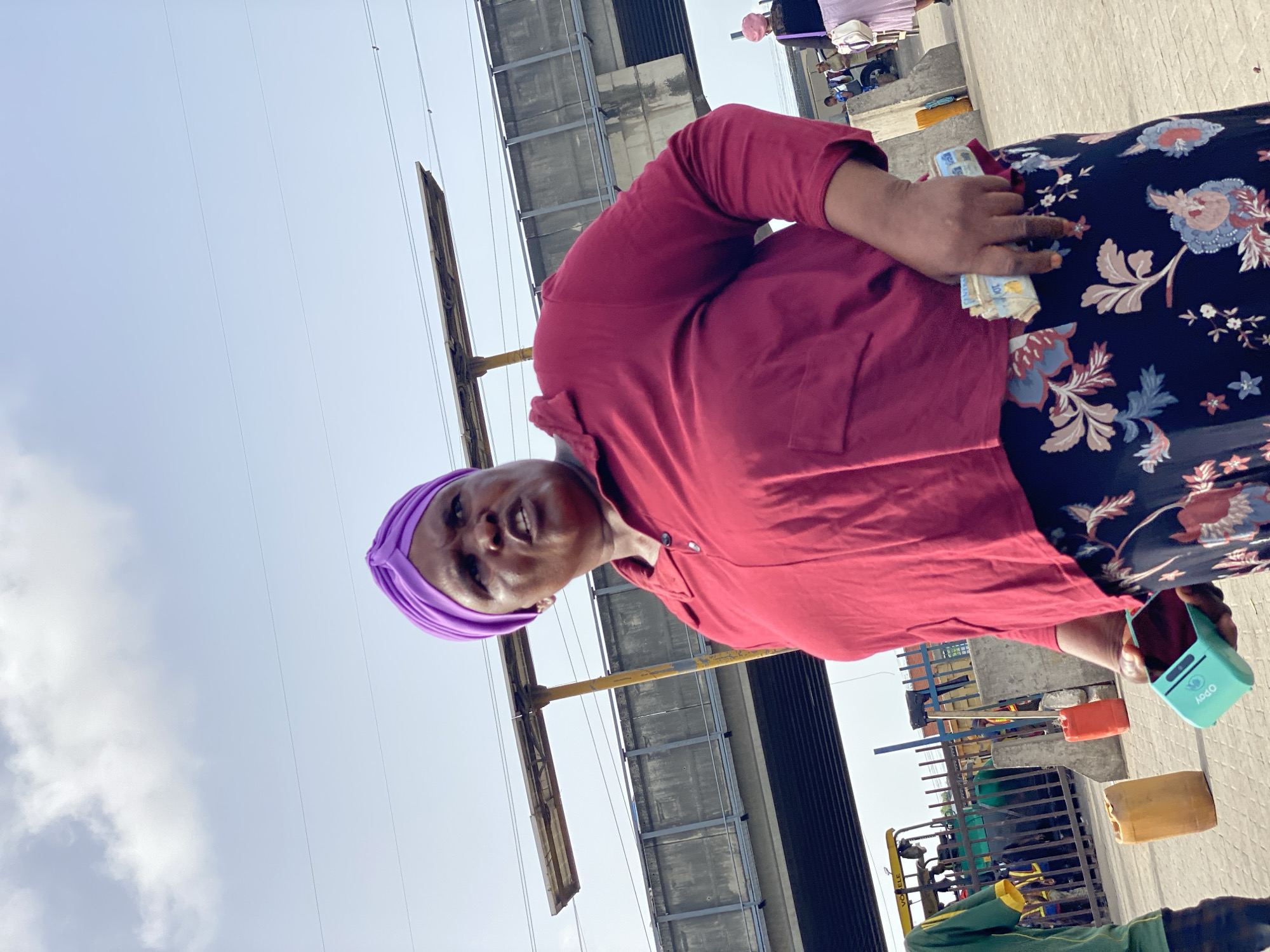 A woman wearing a purple headwrap and a dark red shirt with a floral-patterned skirt stands outside in Ado, Nigeria. She is holding a stack of banknotes in her left hand and a light blue Opay device in her right hand. Behind her is a gray concrete structure with yellow support beams. Other people and objects, including containers and machinery, are visible in the background. The sky is light blue with some clouds.