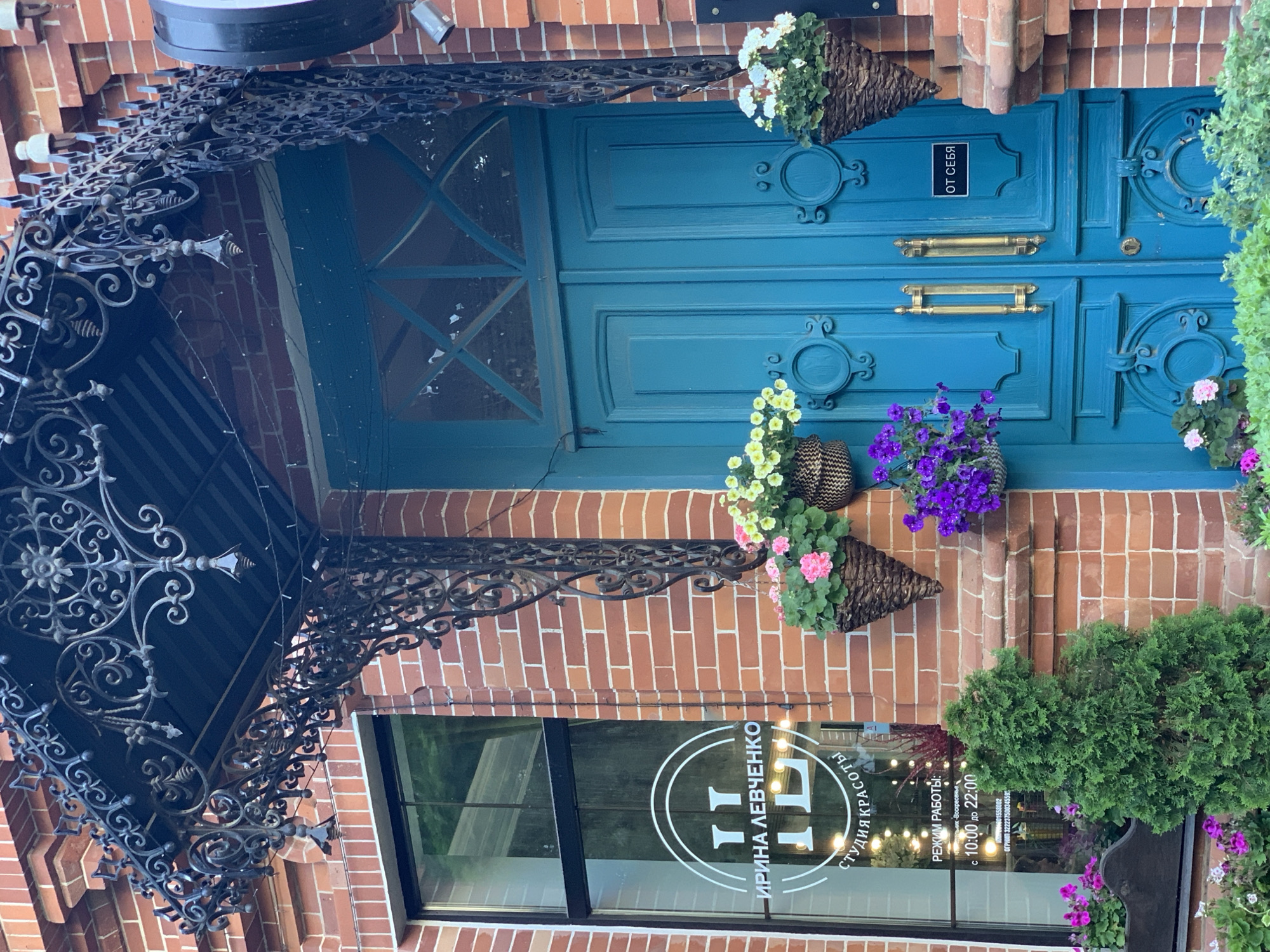 The image shows the exterior of a building in Unknown, Unknown. The facade is brick with a blue double door featuring ornate gold handles and decorative floral baskets hanging on either side. A black wrought iron canopy covers the doorway. Below the door and canopy is a window displaying a white logo with Cyrillic text that reads "ИРИНА ЛЕВЧЕНКО СТУДИЯ КРАСОТЫ," along with business hours. There is greenery on the right side of the door, and some light is visible inside the window.