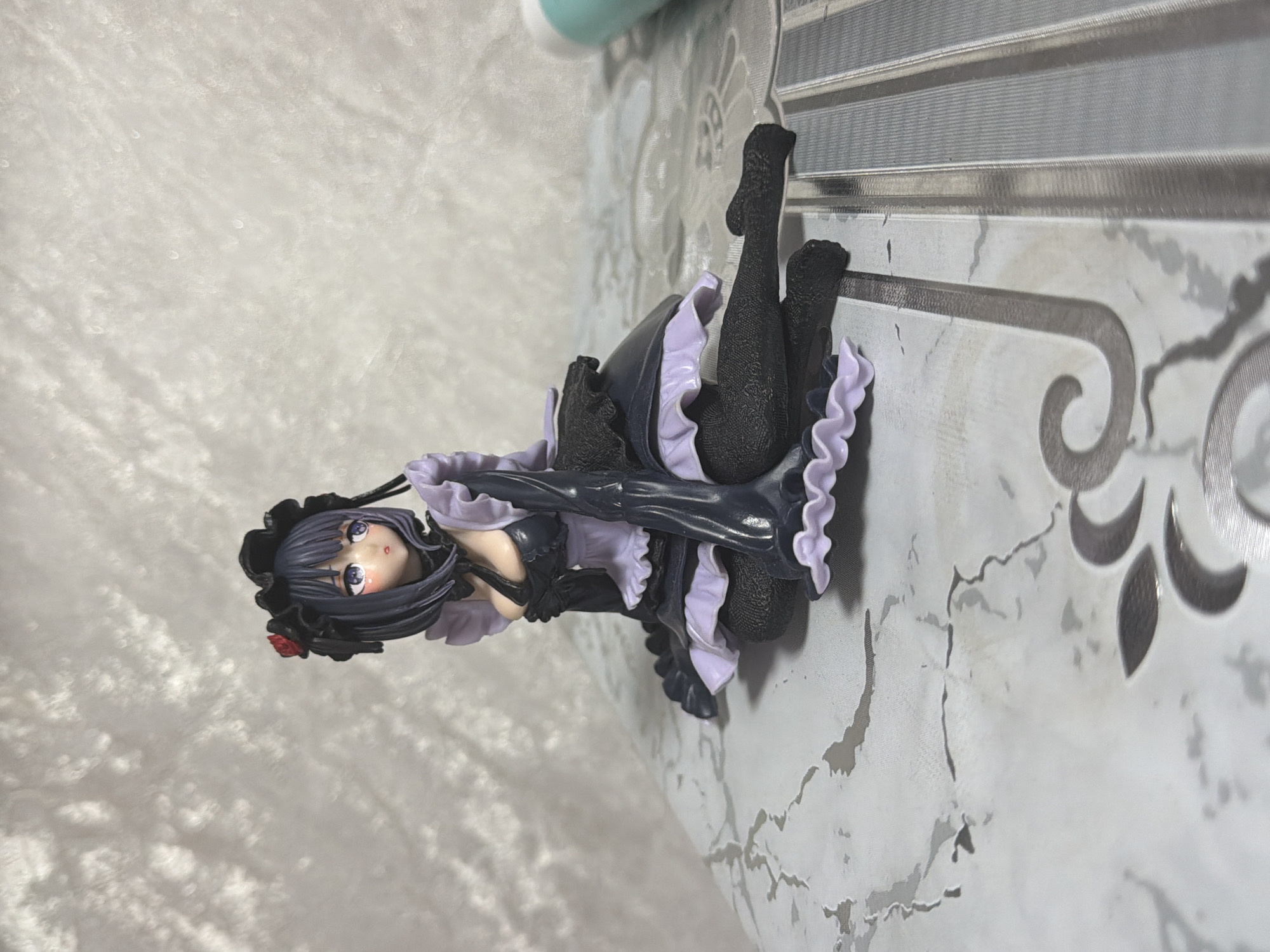 A figurine of a woman in a black and purple maid outfit is displayed on a surface with a marbled pattern. The figurine has dark purple hair adorned with a red rose. She is sitting with her legs crossed and her head tilted to the side, looking to her left. The background is a textured, light-colored surface. The scene is indoors and there are no people present. The location in Saint Petersburg, Russia is not discernible from the image.