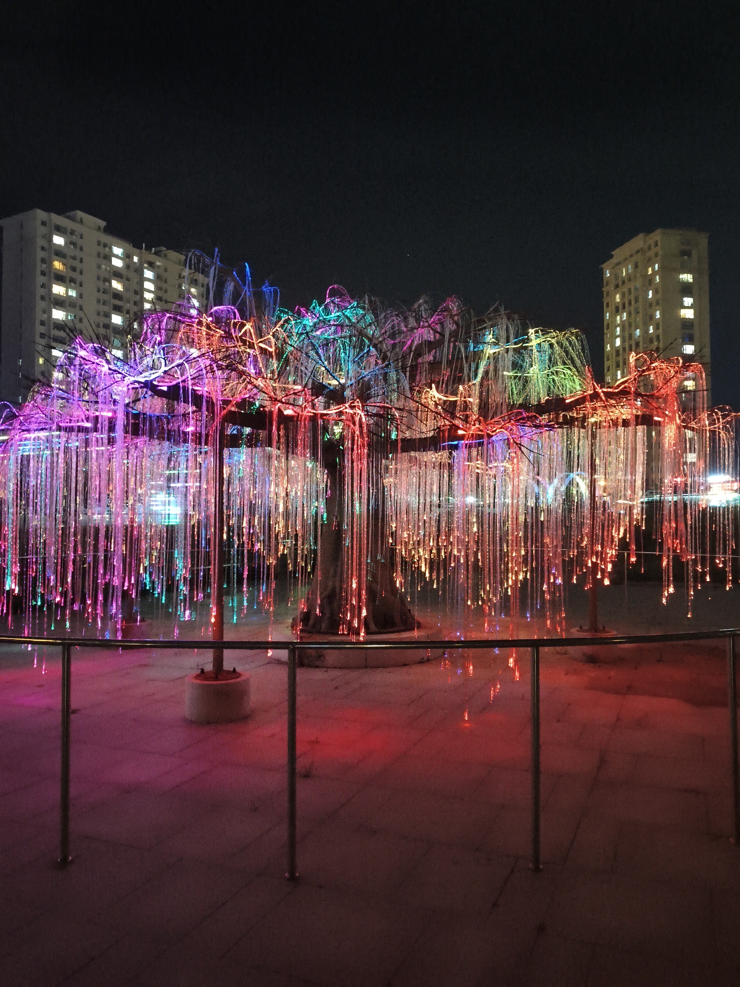 A nighttime outdoor scene features an art installation resembling a weeping willow tree illuminated with numerous colorful LED lights. The tree's branches are adorned with vibrant pink, purple, blue, and green lights, while long, thin, clear strands hang down from the branches, each tipped with a small light that glows in various shades of pink and red. In the background, two tall, multi-story buildings with illuminated windows are visible against the dark night sky. A metal railing in the foreground separates the viewer from the art installation. The ground is paved and reflects the colorful lights from the tree. The location is identified as Jiguan District, China.