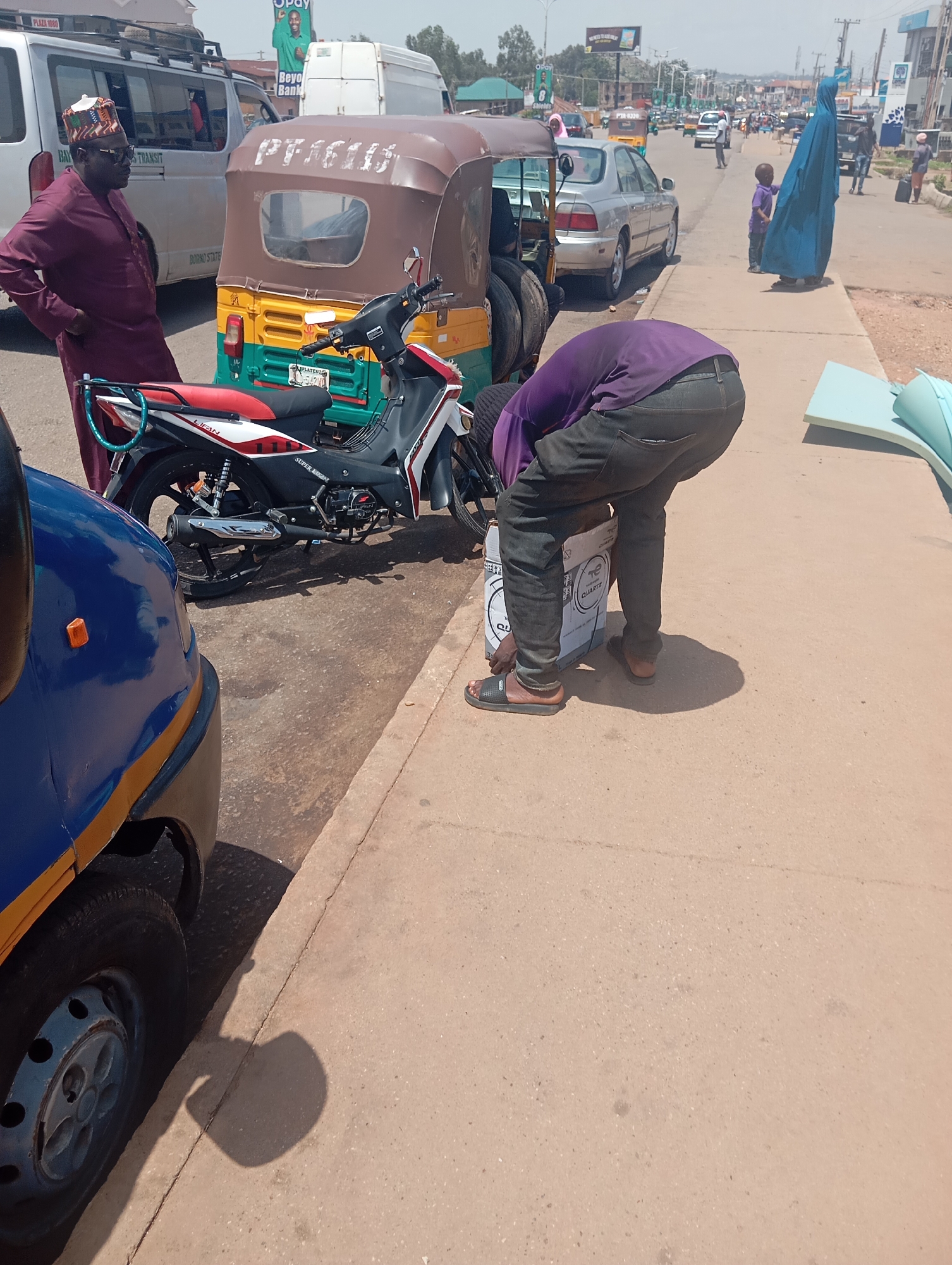 This is a medium-shot, eye-level, outdoor photograph taken on a sunny day in the city of Jos, Nigeria. In the foreground, a blue and yellow auto-rickshaw is partially visible on the left. To the right of the auto-rickshaw, a person wearing a purple shirt and dark pants is bending over to pick up a white cardboard box labeled "QUARTZ". The person is wearing black sandals. Behind the auto-rickshaw is a red, white, and black motorcycle. In the middle ground, a man in a maroon traditional outfit and a matching cap stands with his hands on his hips, looking to the left. Behind him are a white van and a brown and yellow auto-rickshaw. Further in the background, a silver sedan is visible on the road, along with other vehicles and pedestrians. A woman in a blue hijab is walking on the sidewalk with a young child. The scene appears to be a street or busy intersection, possibly a market area. The pavement is light brown, and there are some green trees and buildings in the distance.