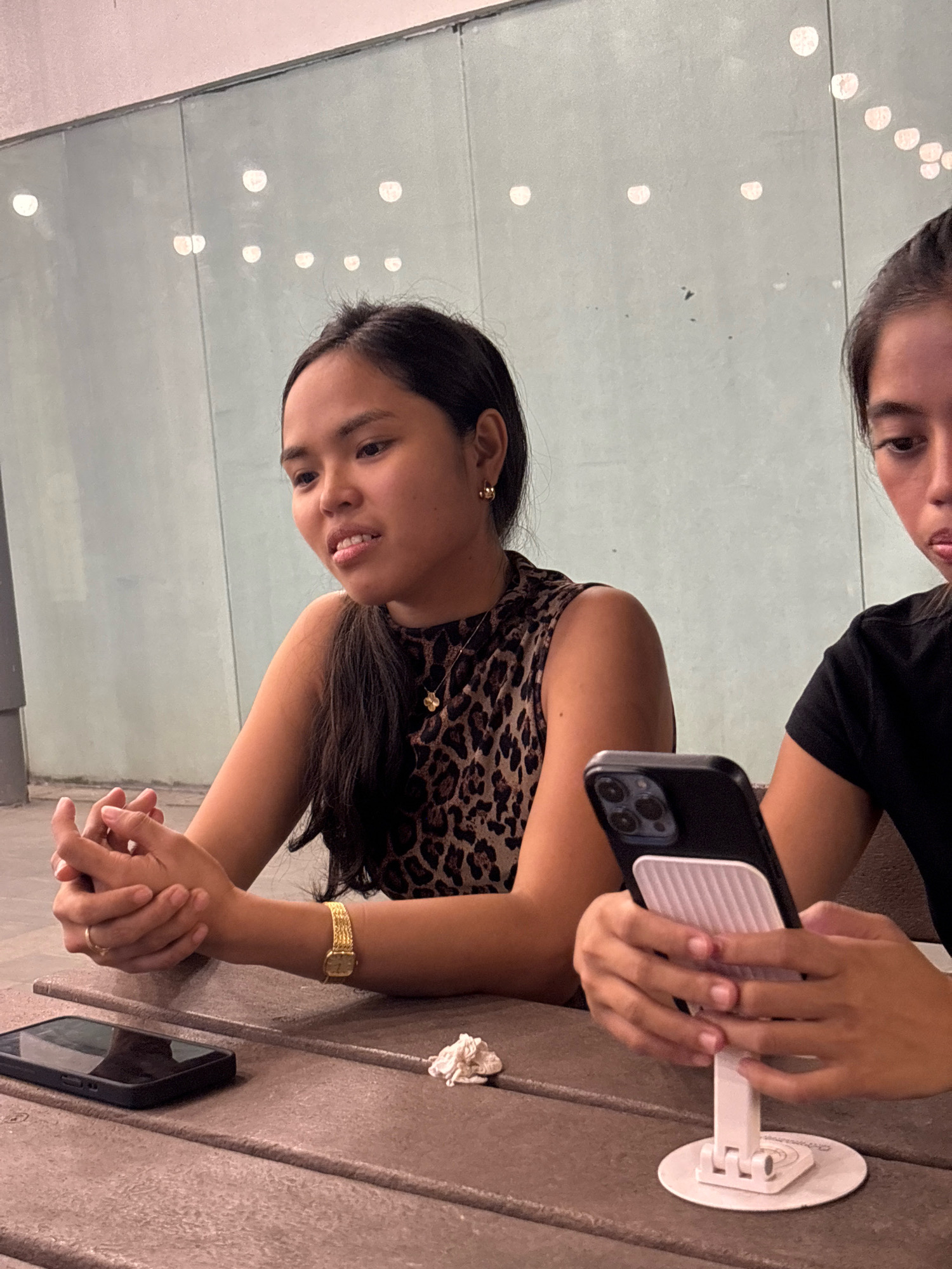 Two young women are seated at a brown picnic table. The woman on the left is wearing a leopard-print sleeveless top and a gold bracelet. Her hands are clasped in front of her. A black smartphone rests on the table to her left. The woman on the right is wearing a black t-shirt and is holding a smartphone on a white stand. The background consists of a light blue wall with circular lights. The scene appears to be an outdoor seating area, possibly at a park or restaurant, in Davao City, Philippines.