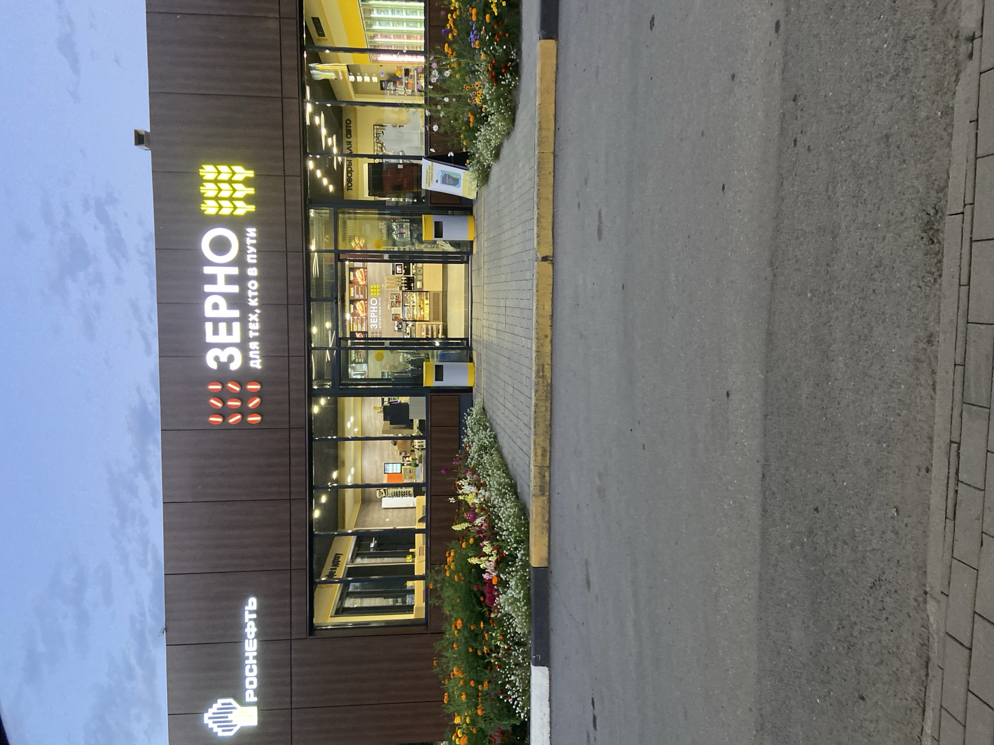 The image shows a gas station and convenience store named "ZERNO" in Slyudyanka, Russia, during dusk. The building has a dark brown exterior with large glass windows. Above the entrance, a prominent sign reads "ZERNO" in large white letters, with a graphic of wheat stalks and the slogan "ДЛЯ ТЕХ, КТО В ПУТИ" (For those on the go) below. To the left of the "ZERNO" sign is the "ROSNEFT" logo, indicating it is a Rosneft branded gas station.

The entrance to the store is open, revealing a brightly lit interior with displays of food items, including pastries and what appear to be sandwiches. Through the windows, customers can be seen inside the store. A flower bed with a variety of colorful flowers is located in front of the building, bordering a paved walkway and the asphalt road. Two grey and yellow trash receptacles are positioned on either side of the entrance. A white A-frame sign with a green graphic is placed near the entrance, advertising a product. The sky above is a pale blue, indicative of twilight. The overall scene depicts a well-lit commercial establishment at the end of the day.