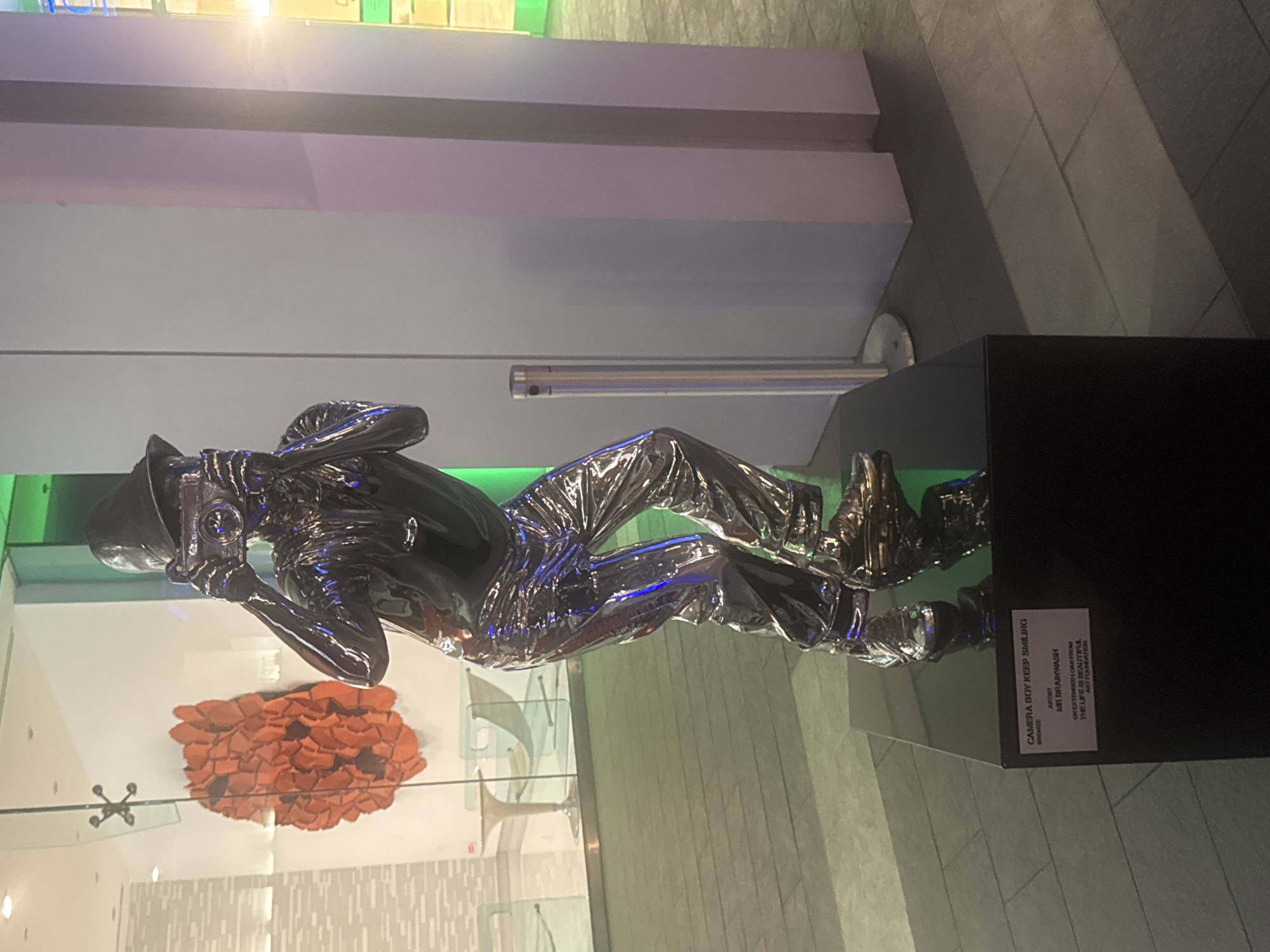 A life-sized, highly polished chrome sculpture of a young person wearing a hat and holding a camera to their face stands on a black pedestal in a modern, urban outdoor setting. The sculpture, titled "CAMERA BOY KEEP SMILING," is attributed to artist MR. BRAINWASH and is on loan from THE LIFE IS BEAUTIFUL ART FOUNDATION. In the background, a wall with abstract orange art is visible through glass, along with modern furniture and a sleek building facade. The sculpture is positioned on a tiled floor, with a silver bollard to its right. The location is West Hollywood, United States of America.