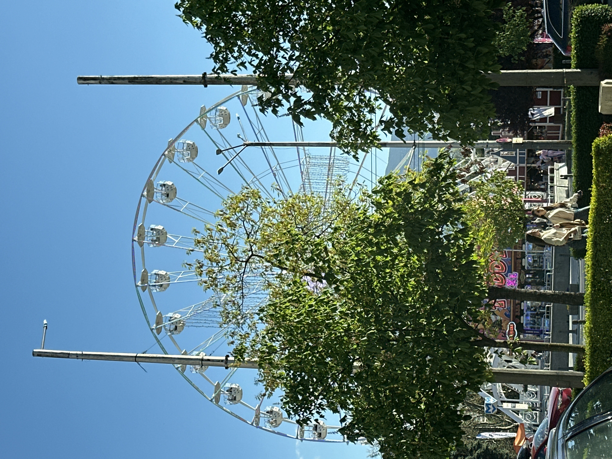 The image captures a large Ferris wheel in what appears to be an urban setting. The wheel is white and situated behind trees. In the background, there are fairground attractions, including buildings or structures with lights, suggesting a carnival or festival. Several people are walking around, indicating activity in the fairground area. Cars are parked along the edge of the scene. The sky is clear and blue. The location appears to be in an urban environment. The image is oriented vertically. Due to the presence of the fairground attractions, the fair is likely in the Borisova gradina park. Therefore, the location in the city is Sofia, Bulgaria, in the area of Borisova gradina.