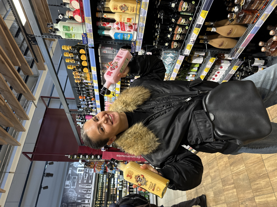 A female subject is depicted standing in the alcohol section of a grocery store or liquor store in Sochi, Russia. She has dark hair pulled back, is smiling, and wears a black bomber jacket with a prominent tan faux fur collar, light blue jeans, and a black cross-body bag. In her left hand, she holds a clear glass bottle containing a yellow liquid, labeled "38 Crema de Tequila Mariachi" with a skull graphic. In her right hand, she holds a clear glass bottle containing a pink liquid, also labeled "38" with a stylized character. Both bottles have black stoppers.
The background features multiple metal shelves stocked with various alcoholic beverages, including Baileys Irish Cream, other cream liqueurs (such as "Bionica"), sparkling wines, and spirits. Price tags in Russian Rubles are visible on the shelves. A vertical sign partially visible between the subject and the products reads "алкогольное" (alcoholic). The ceiling has exposed wooden beams and track lighting. A blurry figure of another individual is partially visible in the far left background. A large black and white wall graphic depicting bottles and glasses is also visible on the left.
