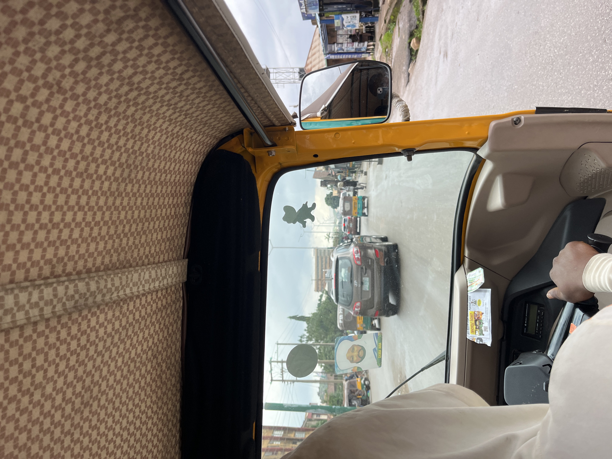 The media file shows the interior view from a yellow tricycle, commonly known as an "okada" or "keke napep" in Nigeria, in Jos. The tricycle is moving on a paved road, likely in an urban or semi-urban area, as indicated by the buildings and other vehicles on the road. The interior of the tricycle features a checkered patterned fabric lining the ceiling and side panels, a common design for such vehicles.
Through the windshield, other vehicles are visible, including cars and other tricycles, suggesting moderate traffic. The sky appears overcast, indicating a cloudy day. A person's hand, presumably the driver's, is visible on the steering wheel, and another person's legs are visible in the foreground, suggesting passengers. A sticker of a man's face with Arabic text, and a "TRICYCLE TICKET" with text in English, are affixed to the inside of the windshield. The presence of a tricycle ticket and the visible landscape are strong indicators of the location being Nigeria.
The overall scene depicts everyday transportation in Jos, Nigeria. The mood appears neutral, capturing a moment of travel. The time of day is difficult to ascertain precisely due to the overcast conditions, but it appears to be daytime.