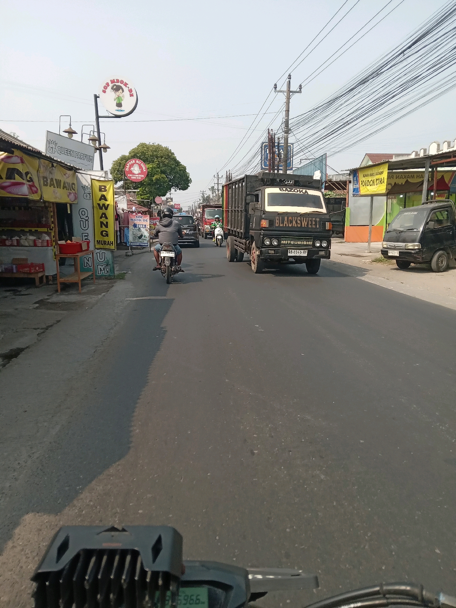 The image shows a street scene in Banguntapan, Indonesia. A black Mitsubishi truck with "BLACKSWEET" written on the side is in the foreground, driving on the right side of the road. The truck has a license plate that reads "AB 8146 RF". To the right of the truck, a small black car is parked. On the left side of the road, there are several stalls with awnings displaying food and merchandise. A person is riding a motorcycle in the middle of the road, heading away from the camera. The sky is clear and blue. The environment appears to be a commercial area with shops and businesses lining the street. There are overhead power lines stretching across the sky.