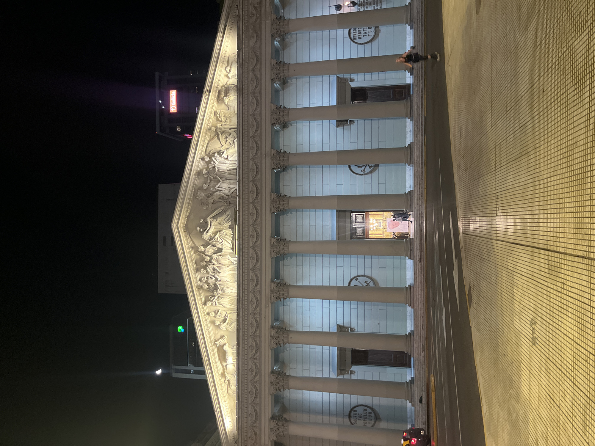 At night, the Metropolitan Cathedral of Buenos Aires, Argentina, presents its facade illuminated by artificial lights. The building, adorned with neoclassical architecture, features a portico of columns and a triangular pediment with a relief of biblical figures. In front of the cathedral, a tiled pedestrian area leads to the entrance. A tall building with red writing on top can be seen behind the cathedral. A person is walking along the facade of the cathedral. At the bottom of the photo is a stoplight that is red.