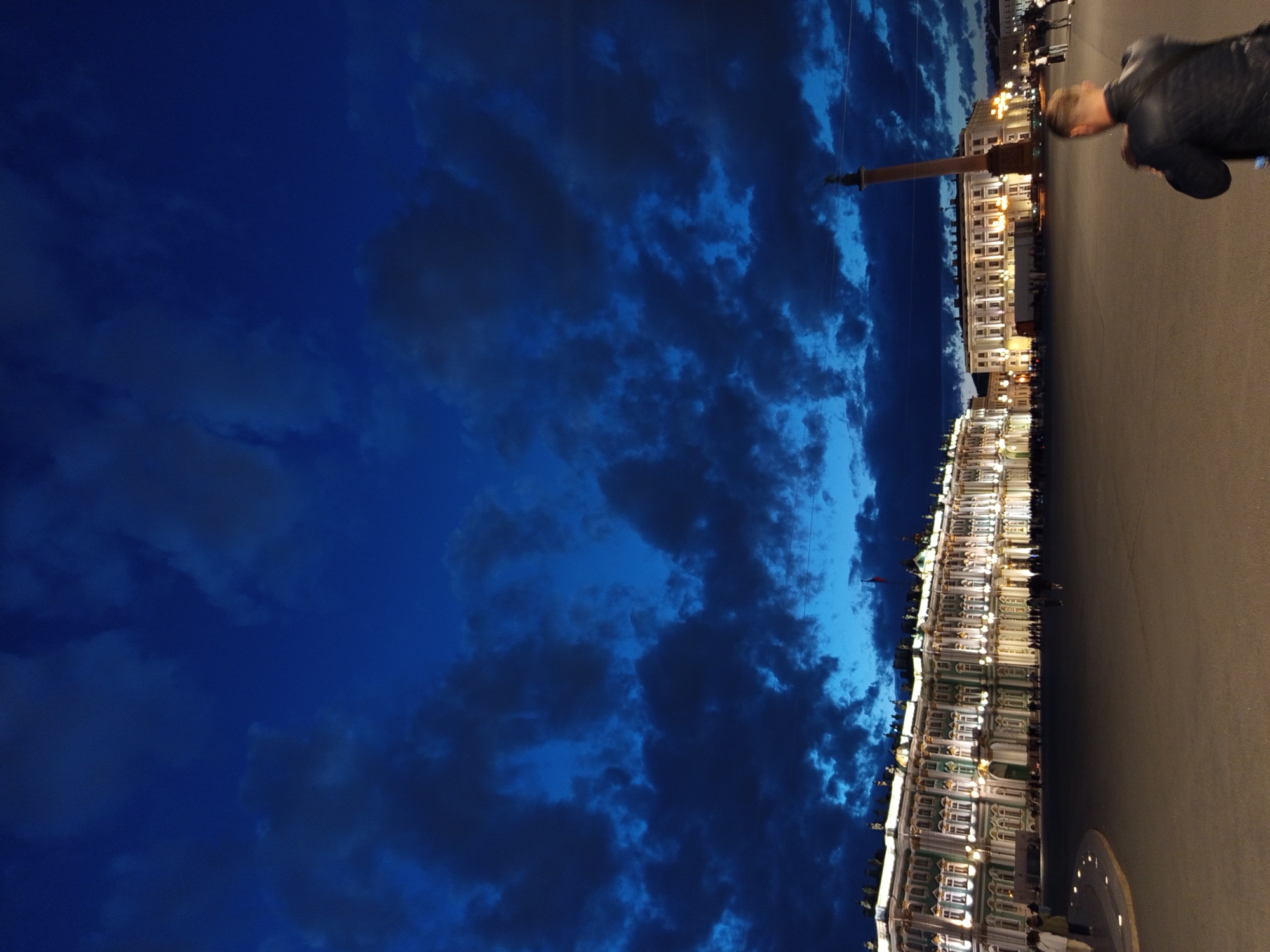 The image depicts Palace Square in Saint Petersburg, Russia, at dusk. A vast, dark blue sky filled with dramatic, swirling clouds dominates the upper portion of the frame. Illuminated buildings, appearing to be the Winter Palace and other governmental structures, line the horizon, their facades aglow with warm, artificial light. A tall, classical column, likely the Alexander Column, stands prominently in the mid-ground, silhouetted against the twilight sky. The square itself is paved, and a few indistinct figures are visible walking across it. In the foreground, a man in a dark jacket is seen from behind, facing away from the camera and towards the illuminated buildings, with his right hand raised to his face, potentially adjusting a camera or taking a photograph. The overall atmosphere is one of grandeur and civic space observed at the transition from day to night.
