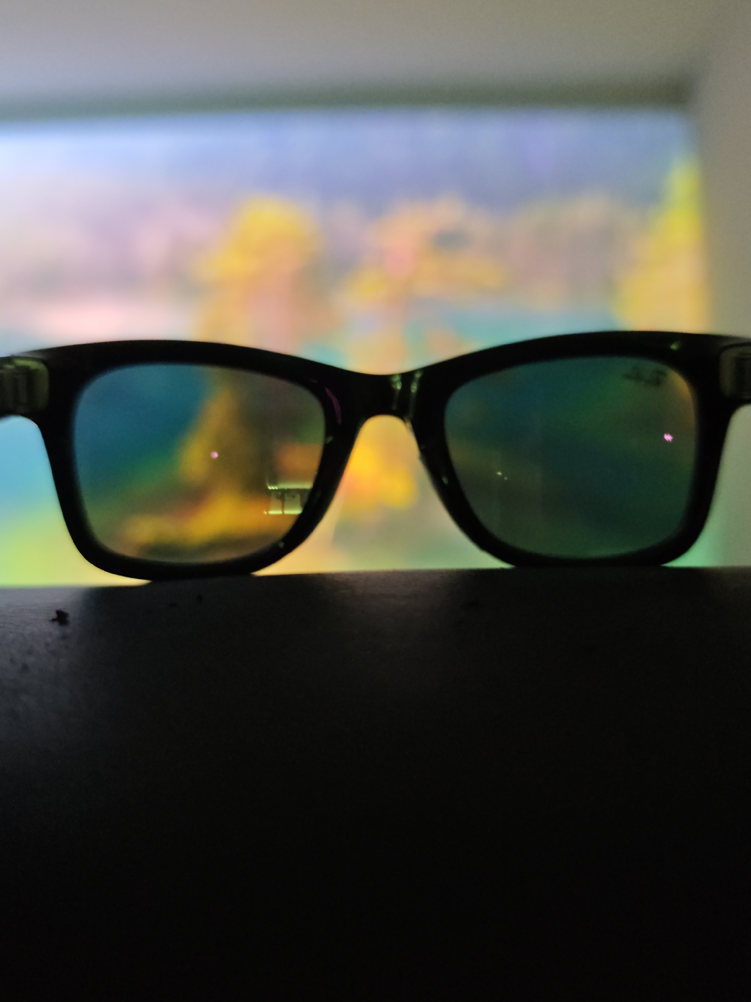 The image features a pair of black-rimmed glasses in the foreground, resting on a dark surface. The lenses of the glasses are reflective, showing a blurred, colorful background that appears to be a projection or a brightly lit wall with abstract patterns of yellow, blue, and pink. The reflection within the glasses also subtly captures some light sources. The overall setting is dark, with the glasses and the surface they rest on silhouetted against the brighter, out-of-focus background. The lighting suggests an indoor environment, possibly a room where a projector or screen is active. There is no discernible text visible in the image. The image does not depict any people or indicate specific weather conditions or a particular time of day. The location context of Calgary, Canada, does not offer any specific visual cues within the image itself.