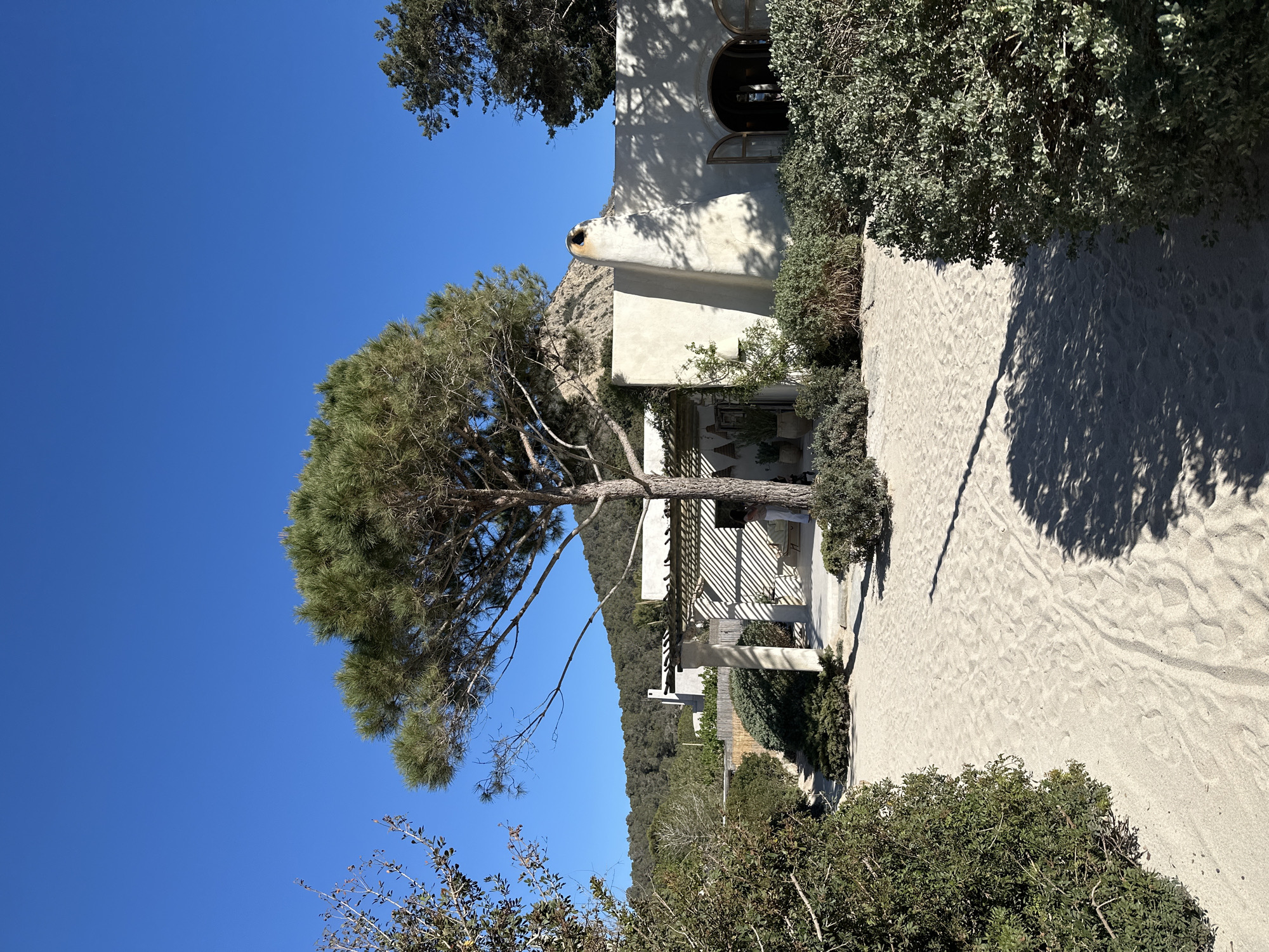 The image shows a white building with an arched window in Sant Josep de sa Talaia, Spain. A large tree stands in front of the building, casting a shadow on a sandy path leading up to it. The sky is clear and blue. The terrain includes low-lying vegetation and a hillside in the background. A pergola structure adjoins the building.