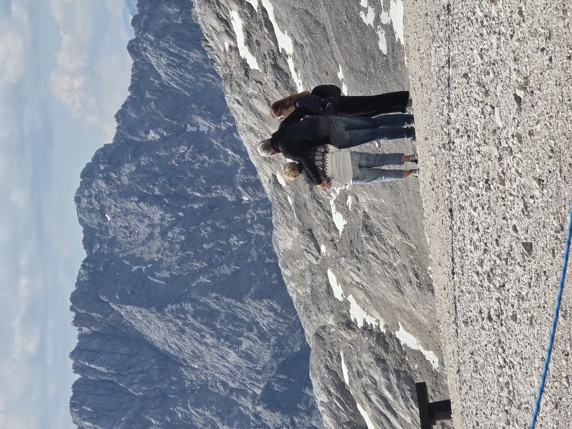 The image depicts a family of three people standing on a rocky, mountainous terrain, with snow patches scattered across the landscape. They are looking out at a majestic mountain range in the background. The people consist of an older man with grey hair, a woman with blonde hair wearing a patterned sweater, and a younger woman with long brown hair. The man has his arm around the woman with blonde hair. The younger woman stands next to them, facing the same direction. In the foreground, a dark bench is partially visible on the left, and a blue rope is stretched across the bottom of the image. The setting appears to be a scenic viewpoint in the Alps, likely at a high altitude. Given the context of Garmisch-Partenkirchen, Germany, this scene is highly likely to be at the Zugspitze, Germany's highest peak, which is accessible by cable car from Garmisch-Partenkirchen and offers such breathtaking mountain vistas.