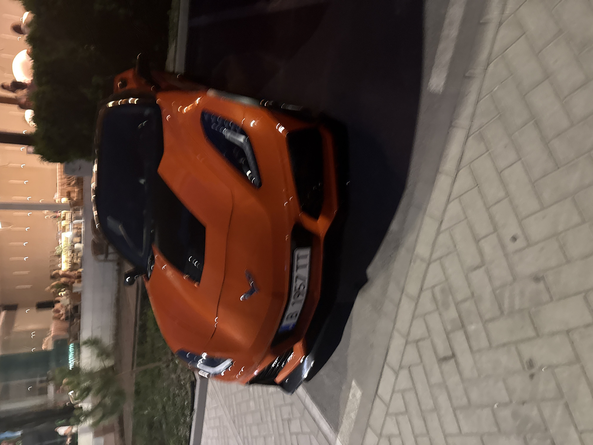 This media file captures an eye-level, slightly angled, full shot of an orange Chevrolet Corvette Stingray parked on a paved sidewalk. The car is the primary subject, with its front end and driver's side visible. The vehicle appears to be a recent model, characterized by its sleek, aggressive design. The license plate is partially visible, displaying the characters "B 1957 TT".
The scene is set outdoors at night, as indicated by the darkness and artificial lighting that creates streaks of light reflecting off the car's surface and nearby foliage. The background is somewhat blurred but suggests an urban or developed area, possibly a promenade or a street lined with buildings or businesses. Hints of greenery and what appear to be outdoor seating areas are discernible, suggesting a place where people gather.
The overall atmosphere is calm and still, with the car appearing to be the only significant object in focus. The lighting suggests that this is a low-light environment, likely illuminated by streetlights or building lights, contributing to the nocturnal setting. The paved ground is composed of rectangular bricks laid in a herringbone pattern. The orientation of the image is rotated, with the car appearing to be on its side, adding a unique perspective to the shot.