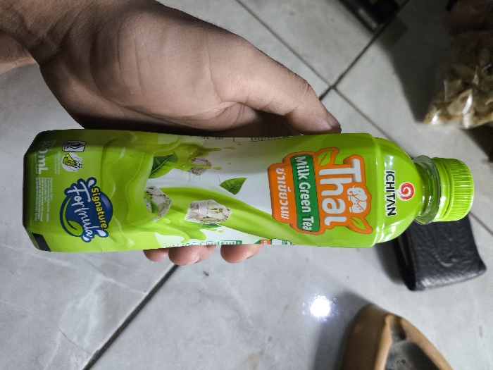 The scene, located in Cibatu, Indonesia, depicts a human hand holding a 310ml green plastic bottle of ICHITAN Thai Milk Green Tea. The bottle features a bright green screw cap and a label primarily in green, white, and orange, displaying the brand "ICHITAN," product name "Thai Milk Green Tea," and "Signature Formula." The hand, appearing to be an adult's, shows visible skin texture and hair on the wrist. The background consists of light grey tiled flooring with dark grout lines. To the immediate right of the bottle, a black object, consistent with a wallet, is partially visible on the floor. Further to the lower right, a light brown shoe or sandal is also partially visible on the tile. A distinct bright light reflection is present on the tiled surface. In the upper background, a blurred, light-colored object, possibly a bag containing snacks, is partially discernible.