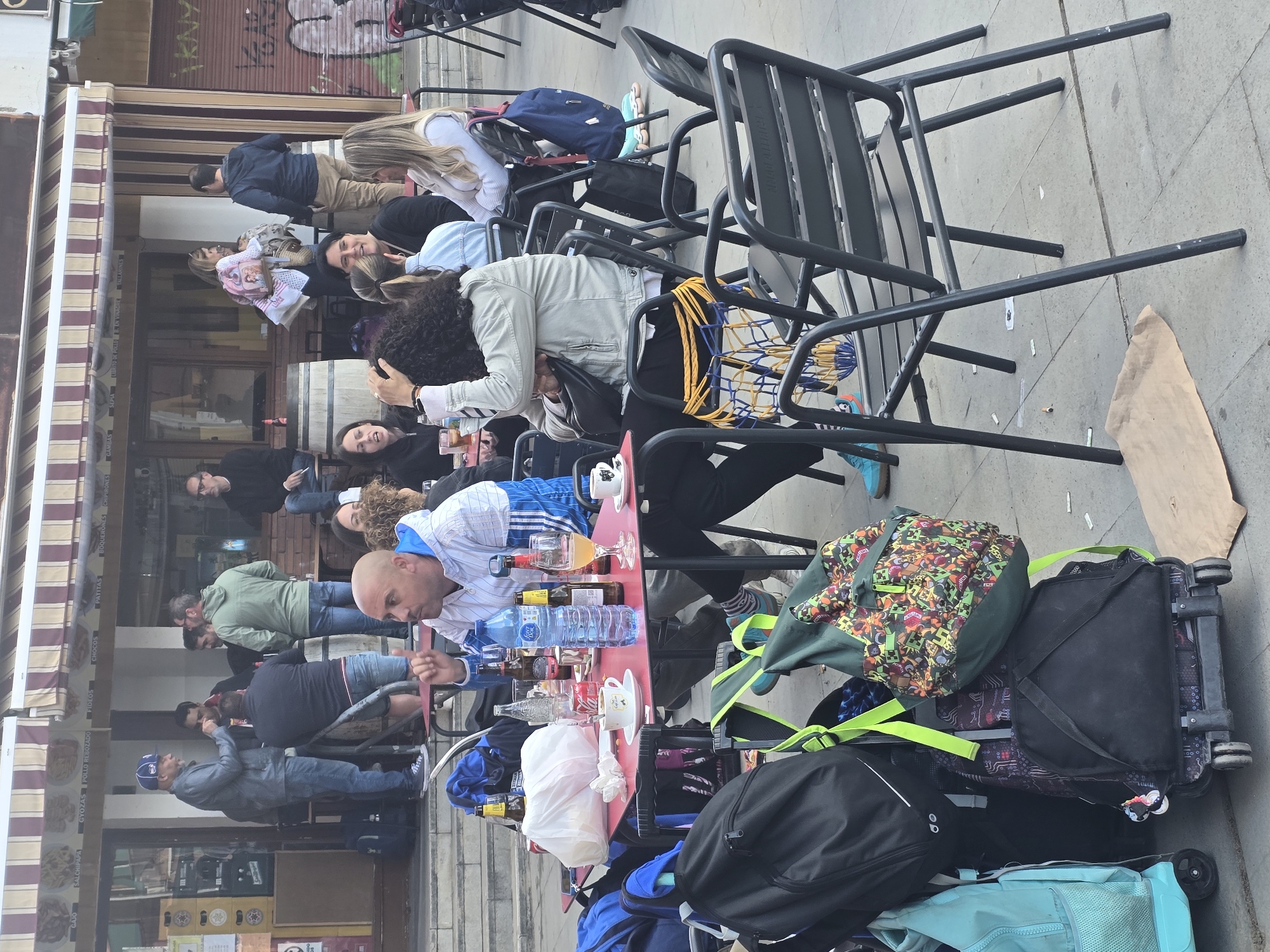 A daytime outdoor scene depicts a group of people seated at a red table outside a building in Cornellà de Llobregat, Spain. Luggage, including rolling suitcases and backpacks, are stacked beside the table, near the chairs. Several people are visible; one man is bald and wears a blue and white striped shirt, another wears a green jacket, and others are in casual clothing. The building has an awning with striped fabric and windows showing the interior. Black metal chairs are stacked nearby. A brown paper bag lies on the pavement. Food and beverages are on the table. The scene suggests a group of travelers pausing at an outdoor cafe or restaurant.