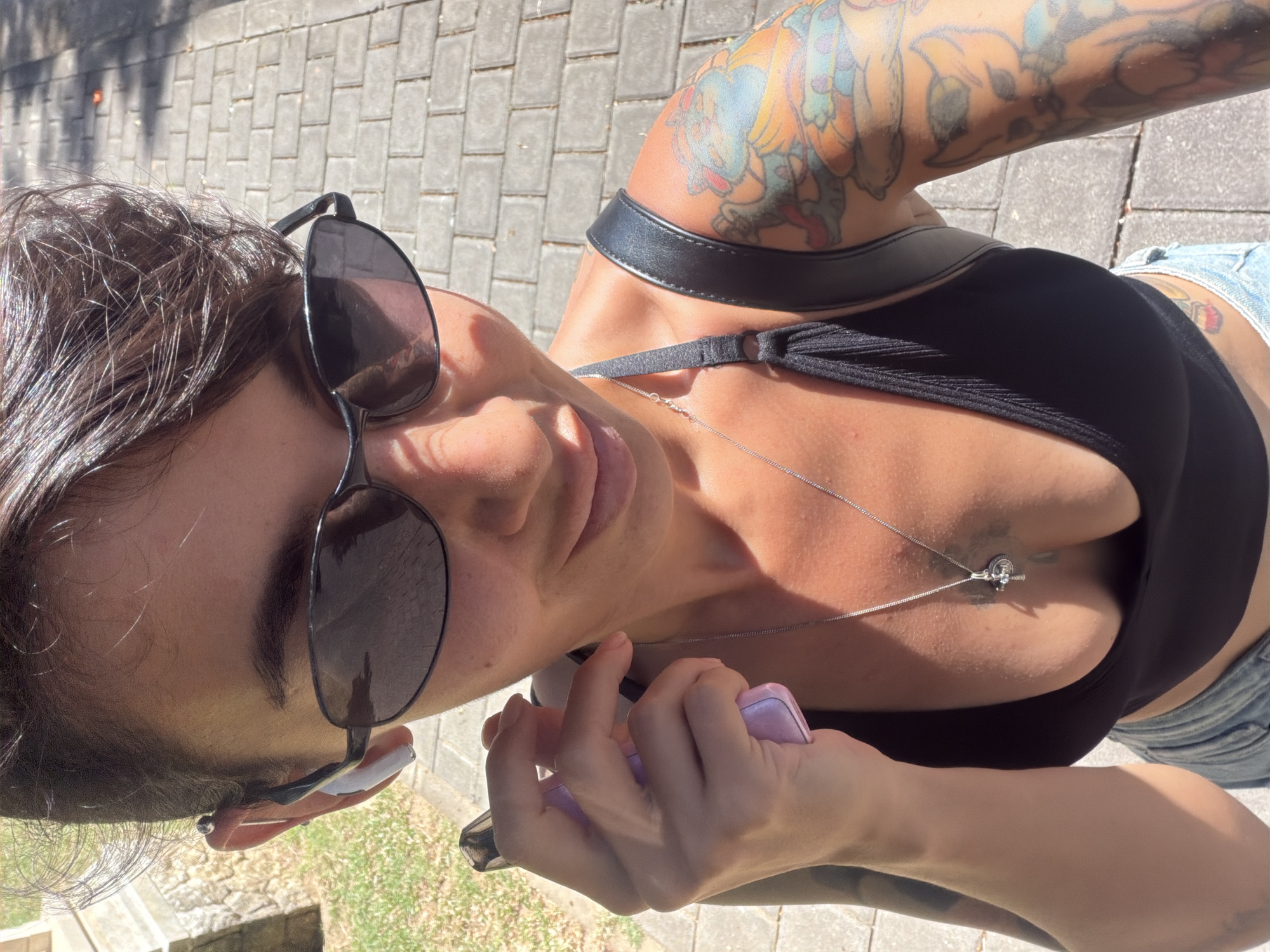 In Kuta Selatan, Indonesia, a woman is captured in a selfie, holding a light purple object to her face. She is wearing dark sunglasses and a black top, with a silver necklace featuring a cross pendant. Her left arm, adorned with colorful tattoos, is visible. The background shows a paved walkway with scattered shadows, suggesting an outdoor setting.
