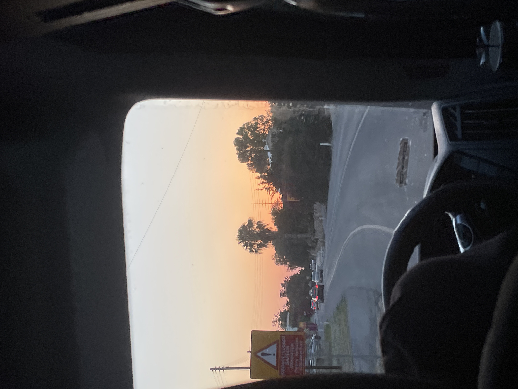 A first-person perspective shot from inside a vehicle shows a road curving to the right at sunset in Acheleia Community, Cyprus. The sky is a gradient of orange and pink. On the left side of the road, a yellow triangular warning sign with a red border displays an exclamation mark and text in red. Trees line the right side of the road, with a large palm tree visible in the middle ground. Several cars are on the road, driving in the same direction. The interior of the vehicle is dark, with a steering wheel and dashboard visible in the foreground.