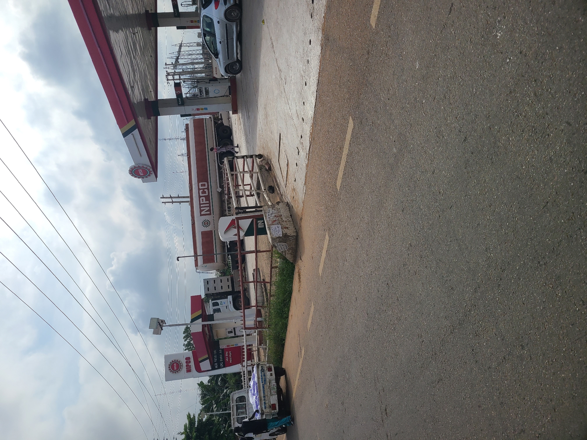 The image captures a roadside scene at a NIPCO fuel station in Karu, Nigeria. A white flatbed truck with blue tarpaulin covering its load is parked on the left side of the frame, with a person in a dark shirt and turquoise pants standing near its rear. In the background, a large NIPCO tanker truck is positioned between two fuel pumps, one of which is visible under a red and white canopy. A silver car is parked to the right of the fuel pumps. The environment appears to be an overcast day, with visible power lines and electrical infrastructure in the background, suggesting an urban or semi-urban setting. The ground surface includes asphalt and concrete, with a paved road in the foreground marked with yellow lines.