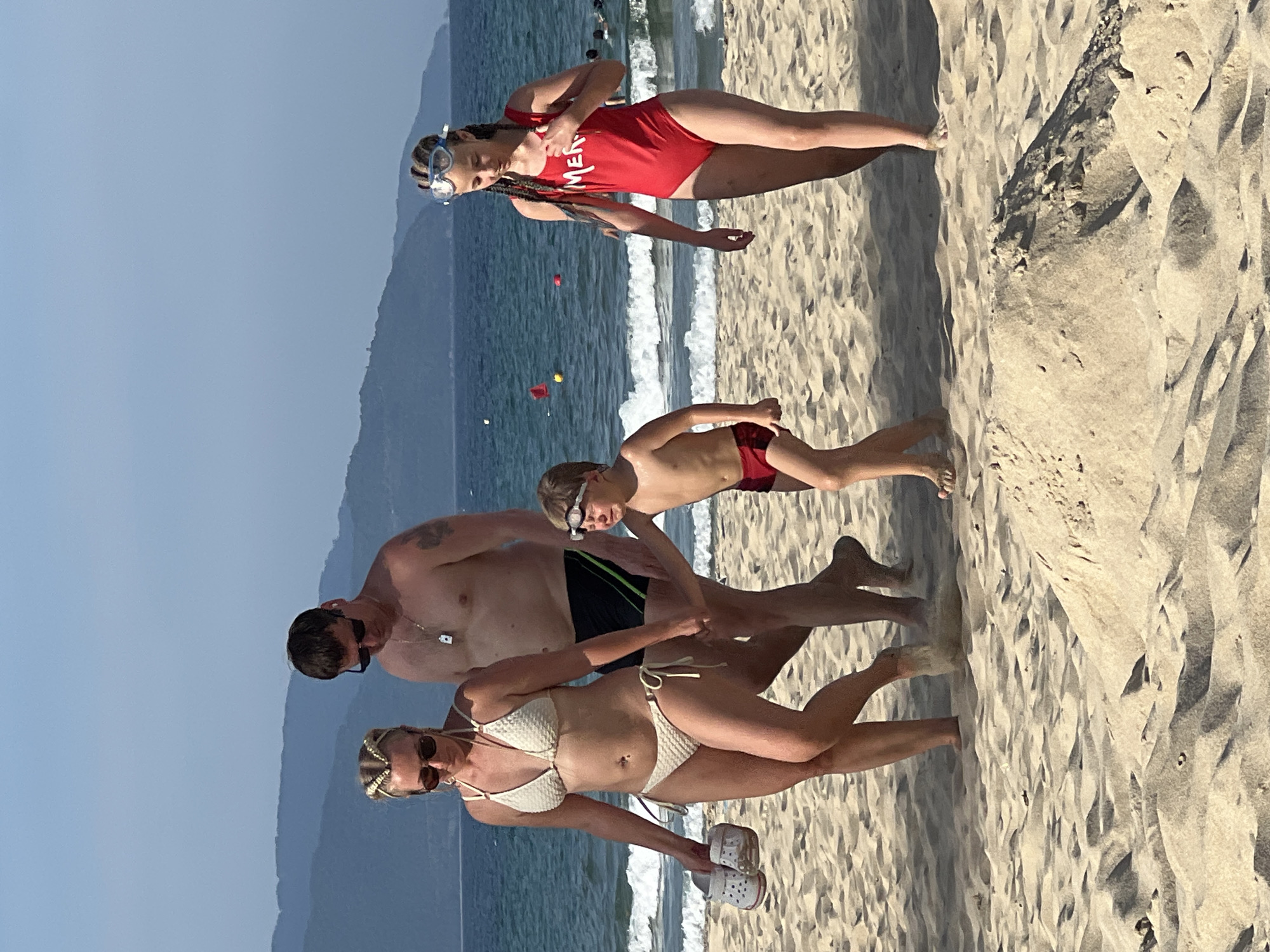 A family is enjoying a day at the beach in Đà Nẵng, Vietnam. The scene is set on a sandy beach with the ocean and waves in the background. The sky is clear and blue, suggesting pleasant weather.
The family consists of four people: two adults, a teenage girl, and a young boy. The teenage girl, positioned at the top of the frame, wears a red swimsuit and goggles, with her hair in braids. She appears to be posing or interacting with someone off-camera. The young boy, in the middle of the frame, is wearing red swim trunks and goggles, looking down and walking. The adult man is to the left, wearing sunglasses and a necklace. To his right is an adult woman wearing a white bikini, sunglasses, and a decorative headband. She is holding a pair of white Crocs. Both adults are looking towards the camera.
The people are dressed in beach attire, indicating they are likely swimming or have just come out of the water. The overall mood seems relaxed and happy, typical of a family vacation. The time of day appears to be daytime, given the bright sunlight casting shadows on the sand. The presence of the ocean and the sandy terrain firmly place the location as a beach. There is visible text on the teenage girl's swimsuit that reads "MENT".