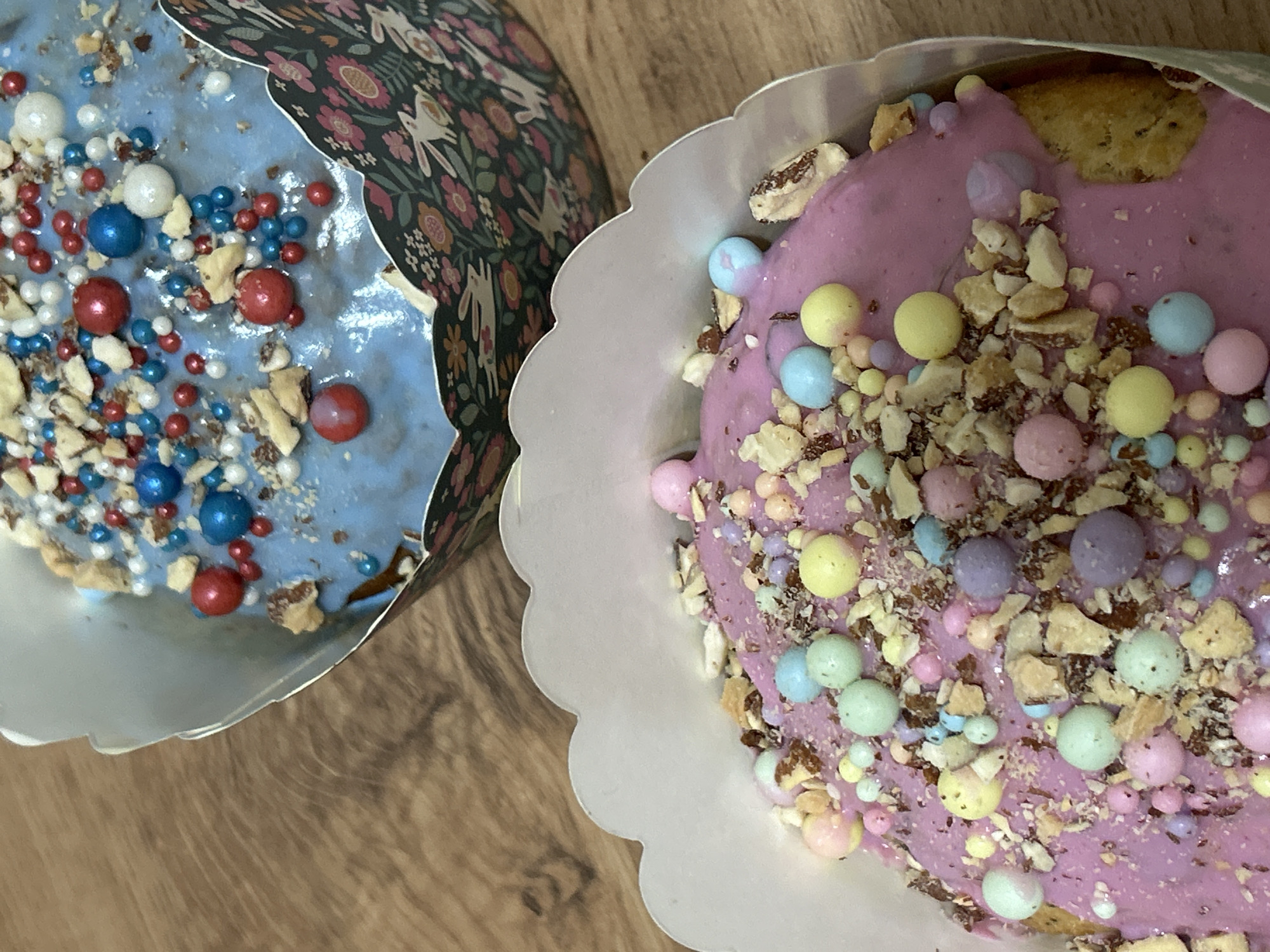 Two Easter cakes are displayed on a wooden surface. The cake on the left has blue icing with red, white, and blue sprinkles and chopped nuts. It is contained in a paper wrapper with a dark floral pattern. The cake on the right has pink icing with pastel-colored sprinkles and chopped nuts. It is in a scalloped white paper wrapper. The photograph was likely taken indoors. There is no visible evidence to confirm the location as Chaikovsky, Russia.