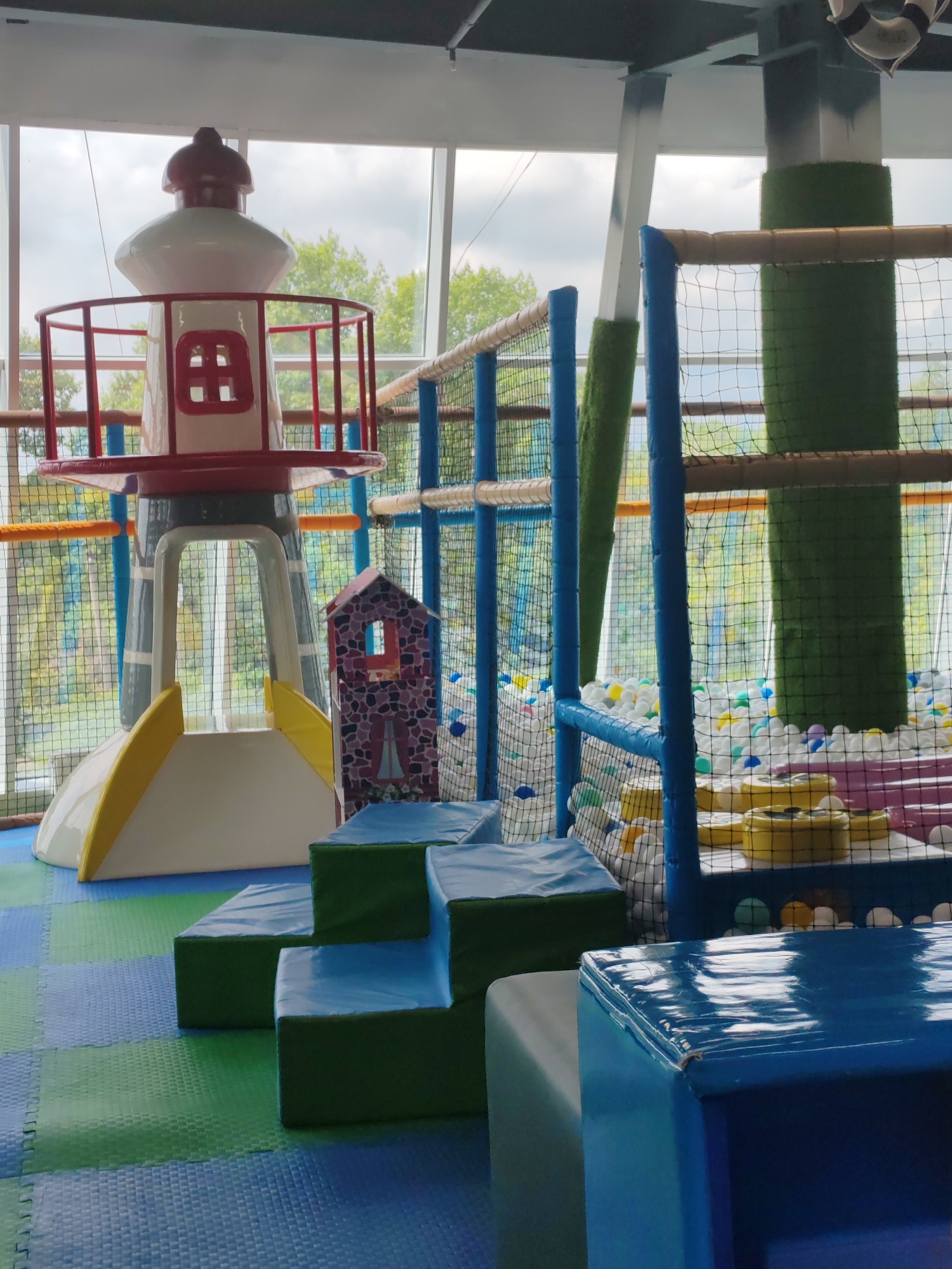 This is a medium-shot, eye-level, indoor photograph of a children's soft play area. The scene is predominantly characterized by blue and green padded flooring tiles. In the foreground, there are blue and green foam blocks arranged in a tiered formation. To the left, a white and red lighthouse-shaped structure with a yellow slide is partially visible. Further back, a ball pit filled with white and colored balls is enclosed by blue netting and wooden poles. Green cylindrical structures, resembling tree trunks or pillars, are interspersed throughout the play area. The background features large windows offering a view of trees and an overcast sky, suggesting an outdoor environment beyond the play space. There are no people visible in the image. The image appears to be taken in the city of Amursk, Russia.