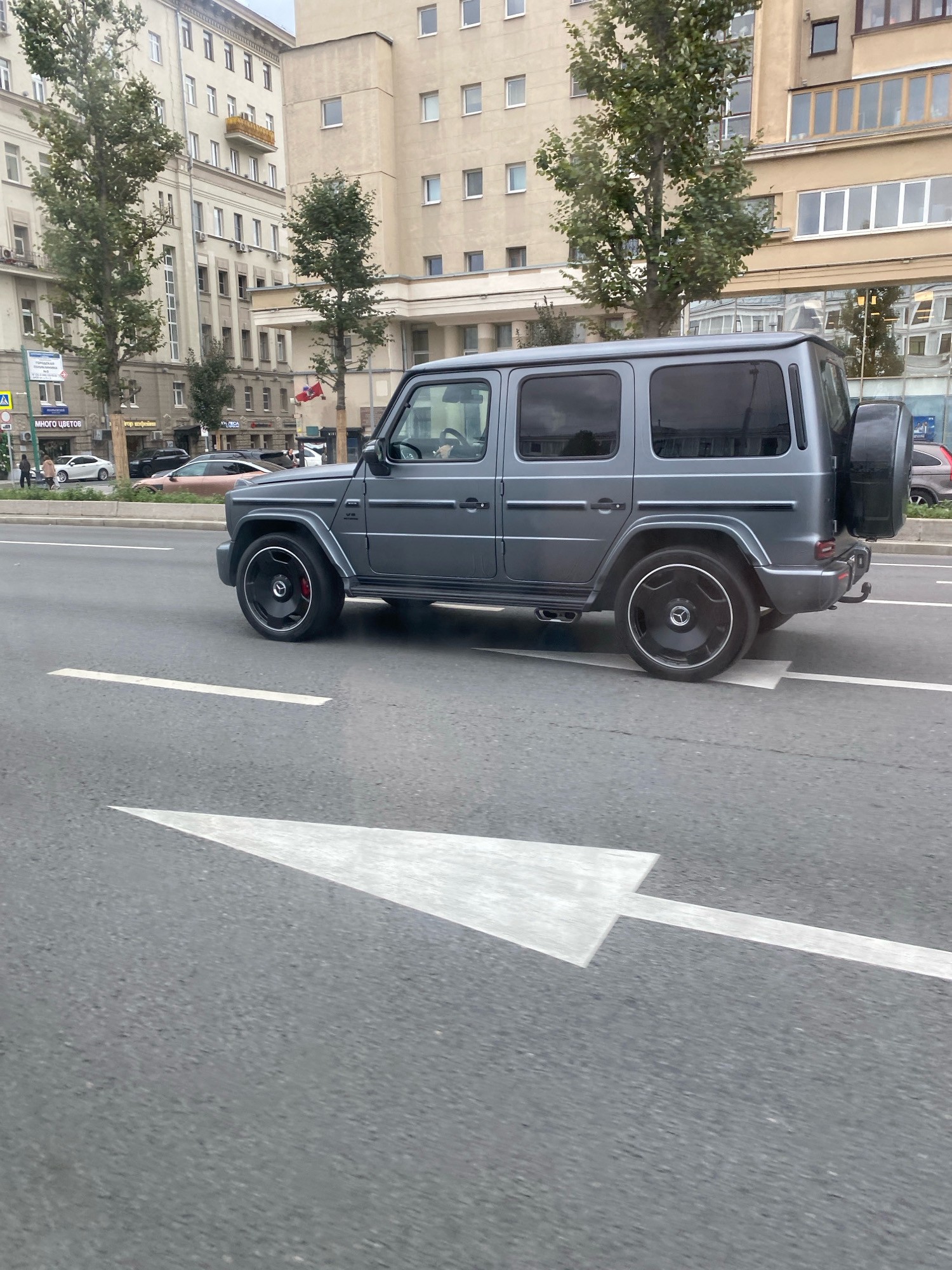A matte gray Mercedes-Benz G-Class SUV is captured in motion on a city street in Moscow, Russia. The vehicle, with tinted windows and a spare tire mounted on the rear, is traveling on the right side of the road, following a white arrow painted on the asphalt, indicating a lane direction. The car is positioned in the foreground, with a portion of its body visible from the front fender to the rear bumper.
Behind the SUV, on the left side of the street, is a multi-story building with numerous windows and balconies. Trees with green foliage are planted along the sidewalk. Several other cars are visible on the road, and some pedestrians are walking on the sidewalk in the distance. The architectural style of the buildings suggests a European city setting, consistent with Moscow. The overall scene depicts a typical urban street view with vehicular and pedestrian traffic under an overcast sky.