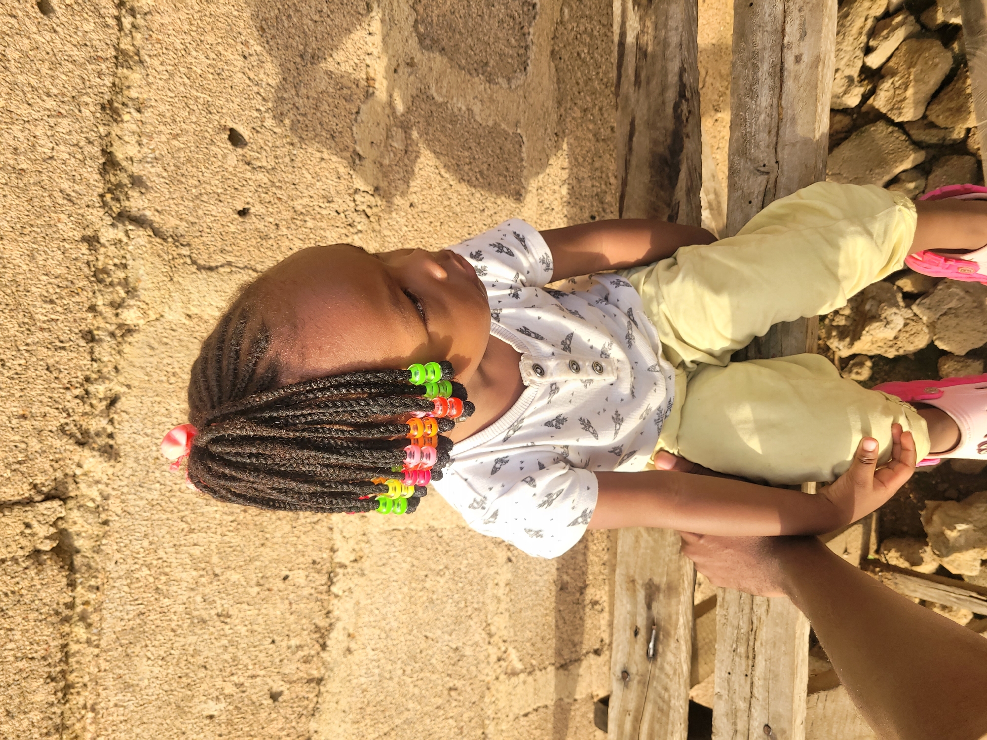 A young child with braided hair adorned with colorful beads sits on a wooden bench in front of a rough, tan-colored wall. The child wears a white t-shirt with a pattern of dark grey birds and yellow pants. The child's right arm is being held by an adult whose arm is visible in the lower left of the frame. The ground is covered with rocks and dirt. The scene appears to be outdoors, possibly in a rural or developing urban area, consistent with locations in Barakin Akawo, Nigeria. Sunlight casts distinct shadows on the wall.
