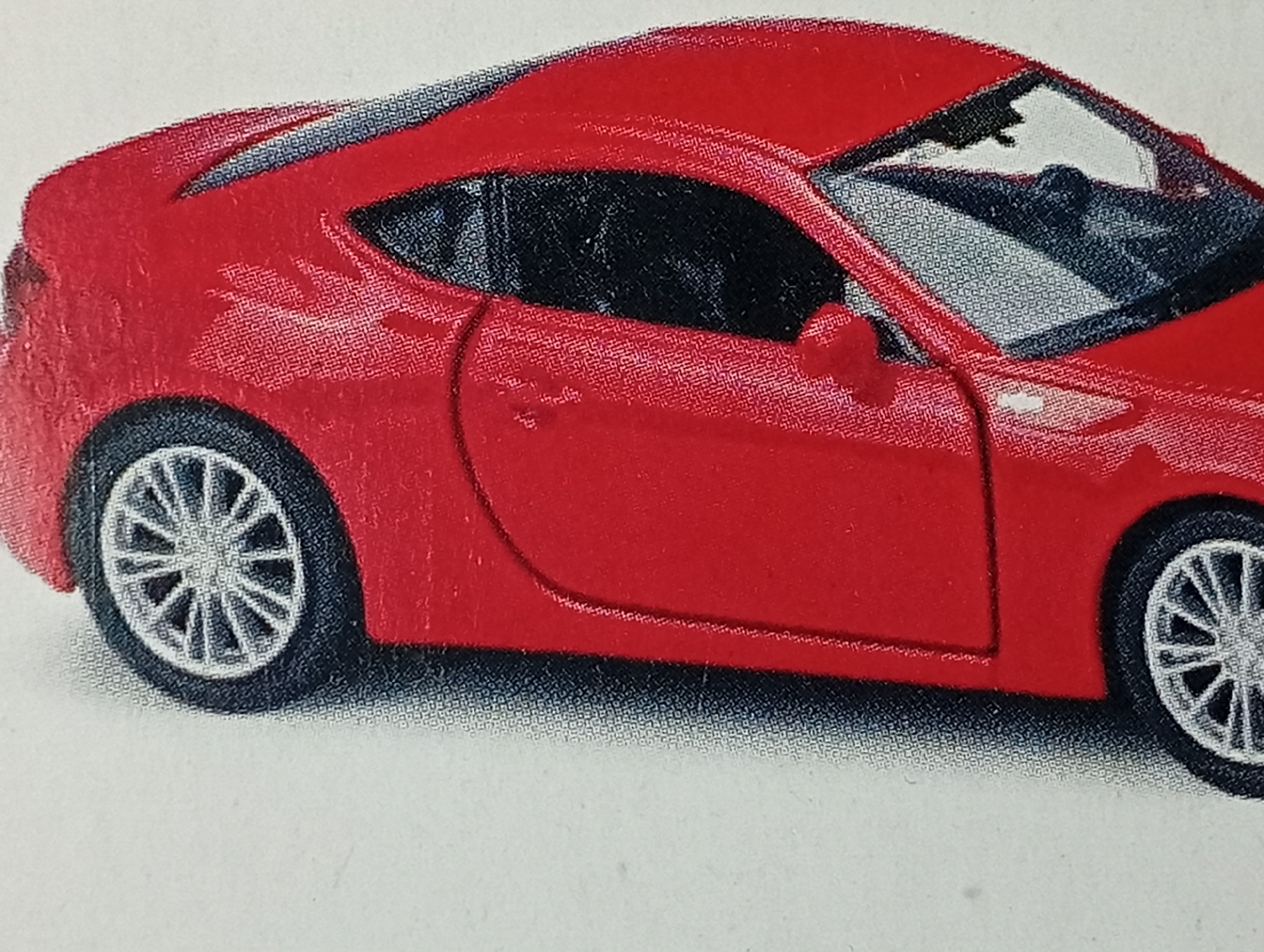 This is a close-up, eye-level photograph of the side of a red toy car. The car is a sports car model with black tinted windows and silver rims on its wheels. The background is a plain white surface. There are no people, other objects, or environmental features visible in the image. The image does not provide any information about the city of Lhokseumawe, Indonesia.