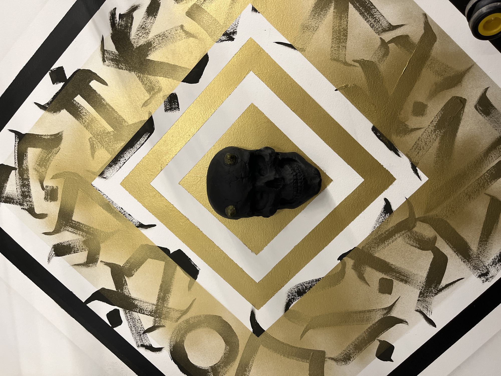The image depicts an abstract artwork, featuring a black skull centrally positioned on a geometric design composed of alternating gold and white square frames. Surrounding the central frame is a layer of gold brushstrokes resembling calligraphy. The artwork is further bordered by a black square frame. The location, Chaikovsky, Russia, cannot be verified from the image.