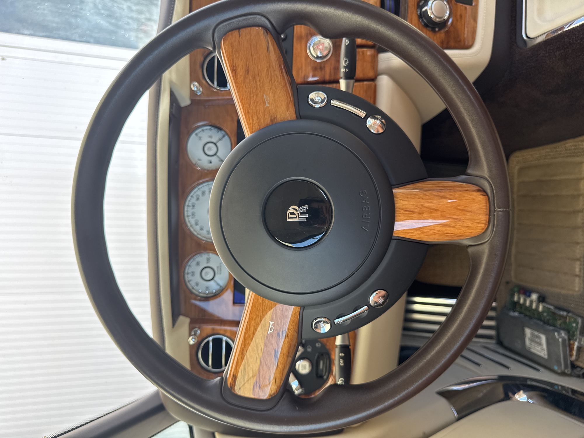 This is a close-up, eye-level shot of the interior of a luxury vehicle, likely a Rolls-Royce, focusing on the steering wheel and dashboard. The steering wheel is dark brown leather with polished wood accents and a black center hub featuring the Rolls-Royce emblem and "AIRBAG" text. On the dashboard, visible in the background, are circular gauges and wood trim. The image is taken indoors, possibly in a garage or showroom, given the clean and uncluttered environment. There are no people or animals visible in the image. The lighting suggests it is daytime, possibly with artificial light contributing. The location of Dub, Montenegro is not discernible from the visual information provided.