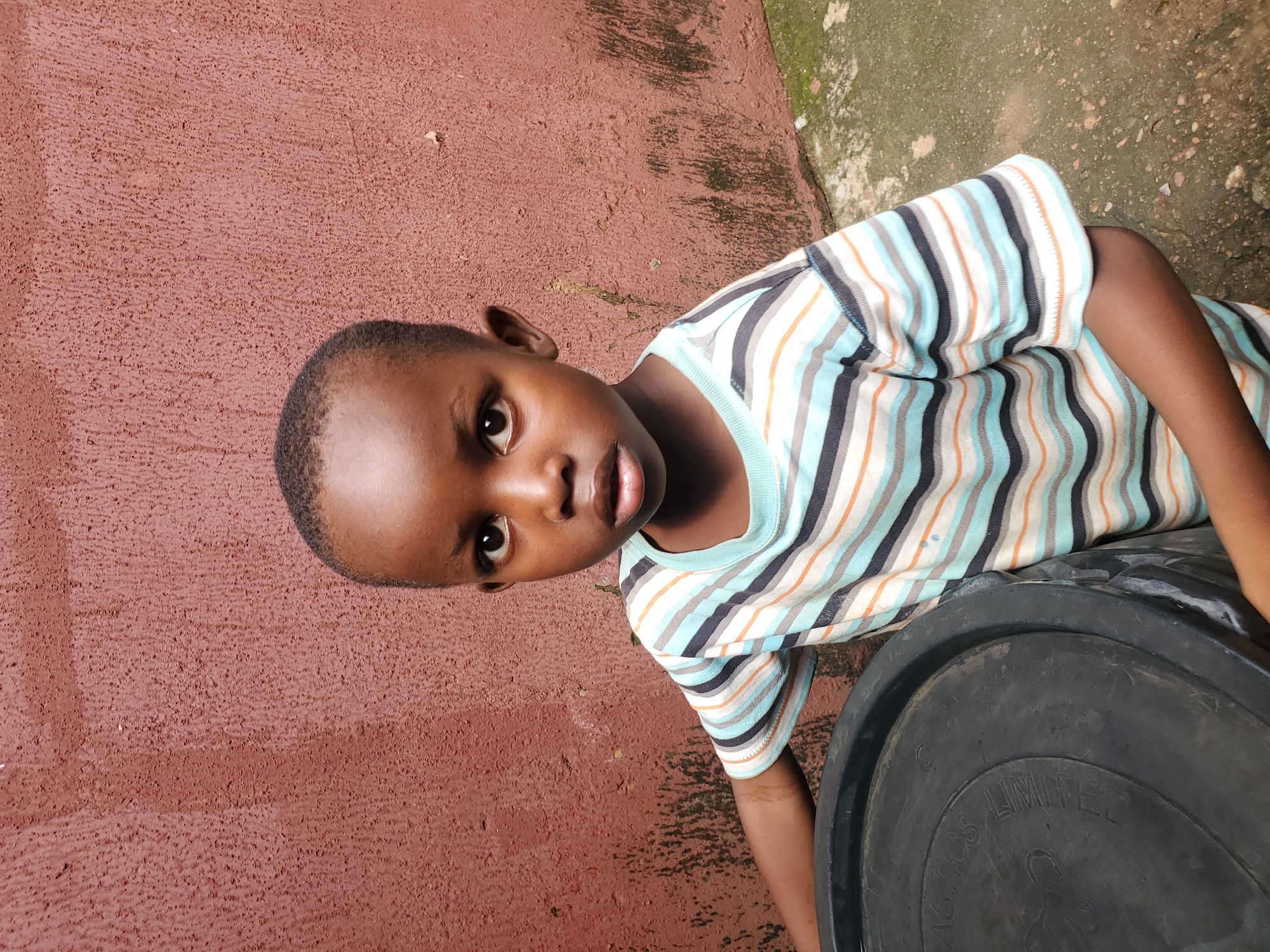 A young boy with dark skin and short hair is looking directly at the camera. He is wearing a striped t-shirt with alternating bands of light blue, white, and orange. His left arm is resting on a large black object, possibly a bucket or drum, which has some text visible on its surface. The boy's expression is neutral.
The background consists of a textured, reddish-brown wall with some patches of green and gray, suggesting it is outdoors. The ground surface is visible in the upper right corner, appearing to be a rough, earthy or concrete texture with small stones. The lighting suggests it is daytime, possibly mid-morning or afternoon. The overall scene is casual and appears to be a snapshot of everyday life.
The visible text on the black object reads "PLASTICS LIMITED". The location context is Jos, Nigeria.
